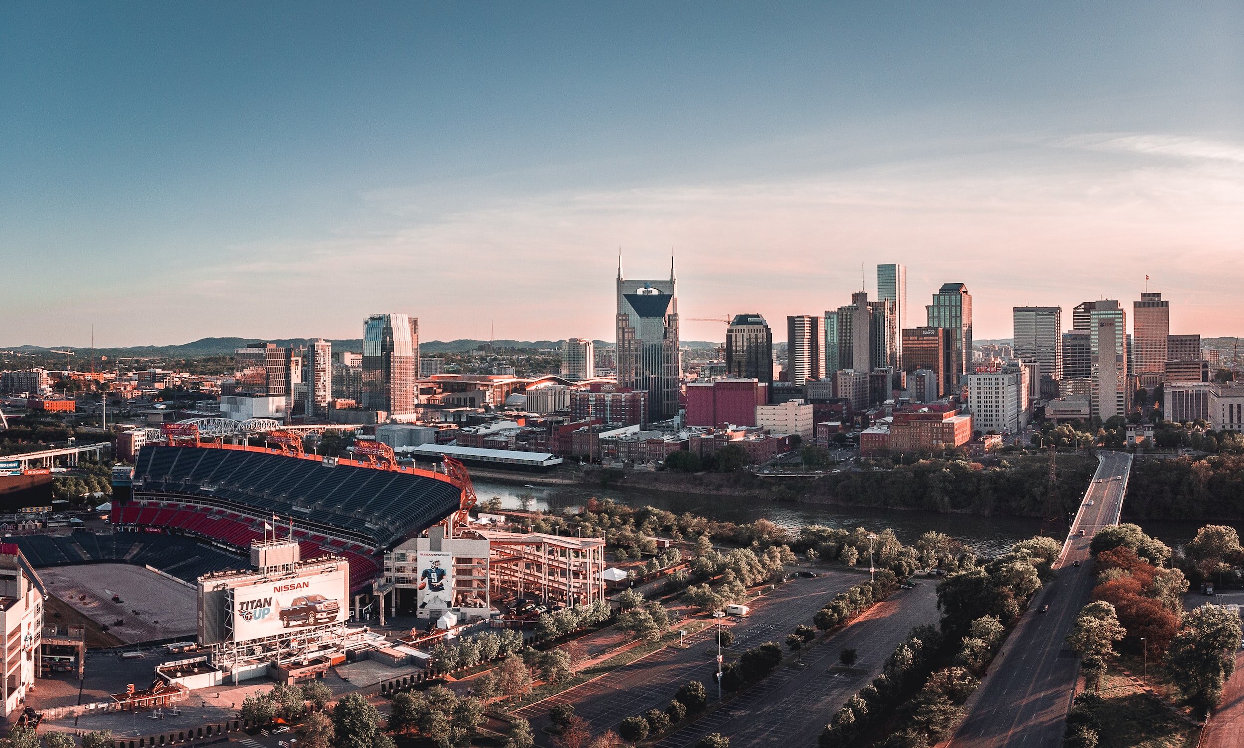 Aerial view of a city skyline at sunset with tall skyscrapers, a river, a bridge, a stadium, and parking lots.