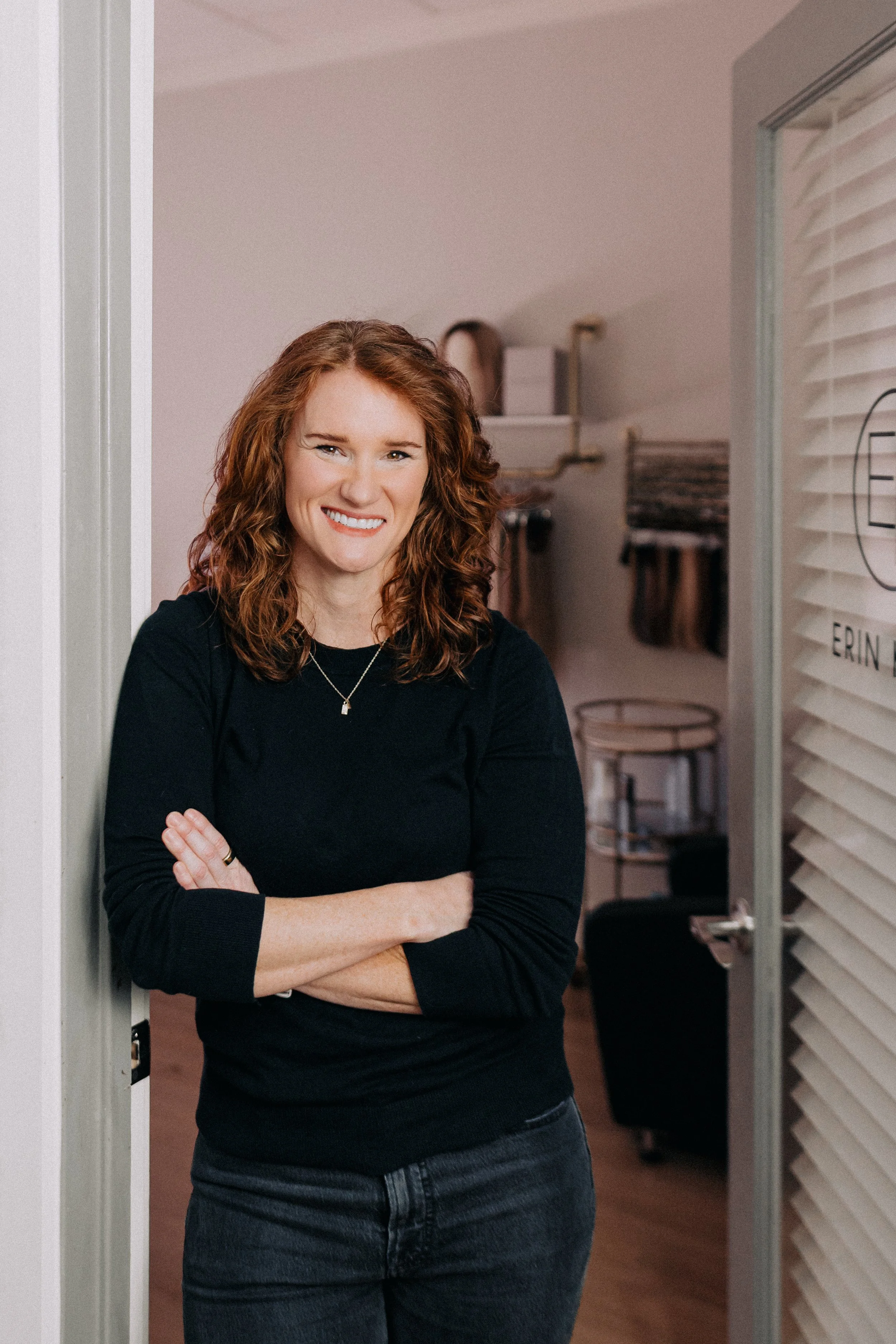 Young woman with curly red hair smiling and crossing her arms, standing in a room with white walls and partial view of a hallway.