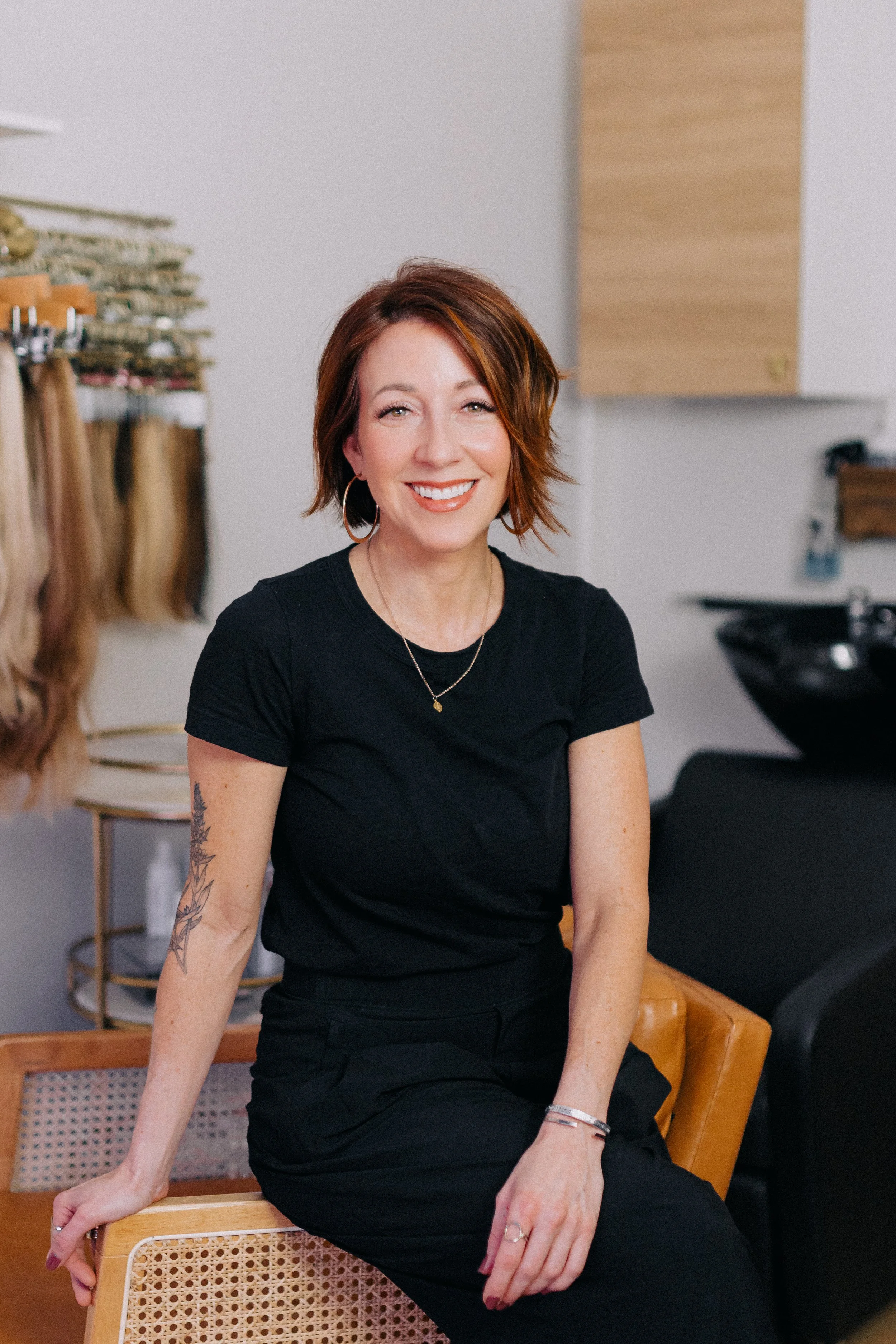 A smiling woman with short red hair and hoop earrings, wearing a black t-shirt and black pants, sitting on a wooden chair in a room with hair swatches and a hairdryer in the background.