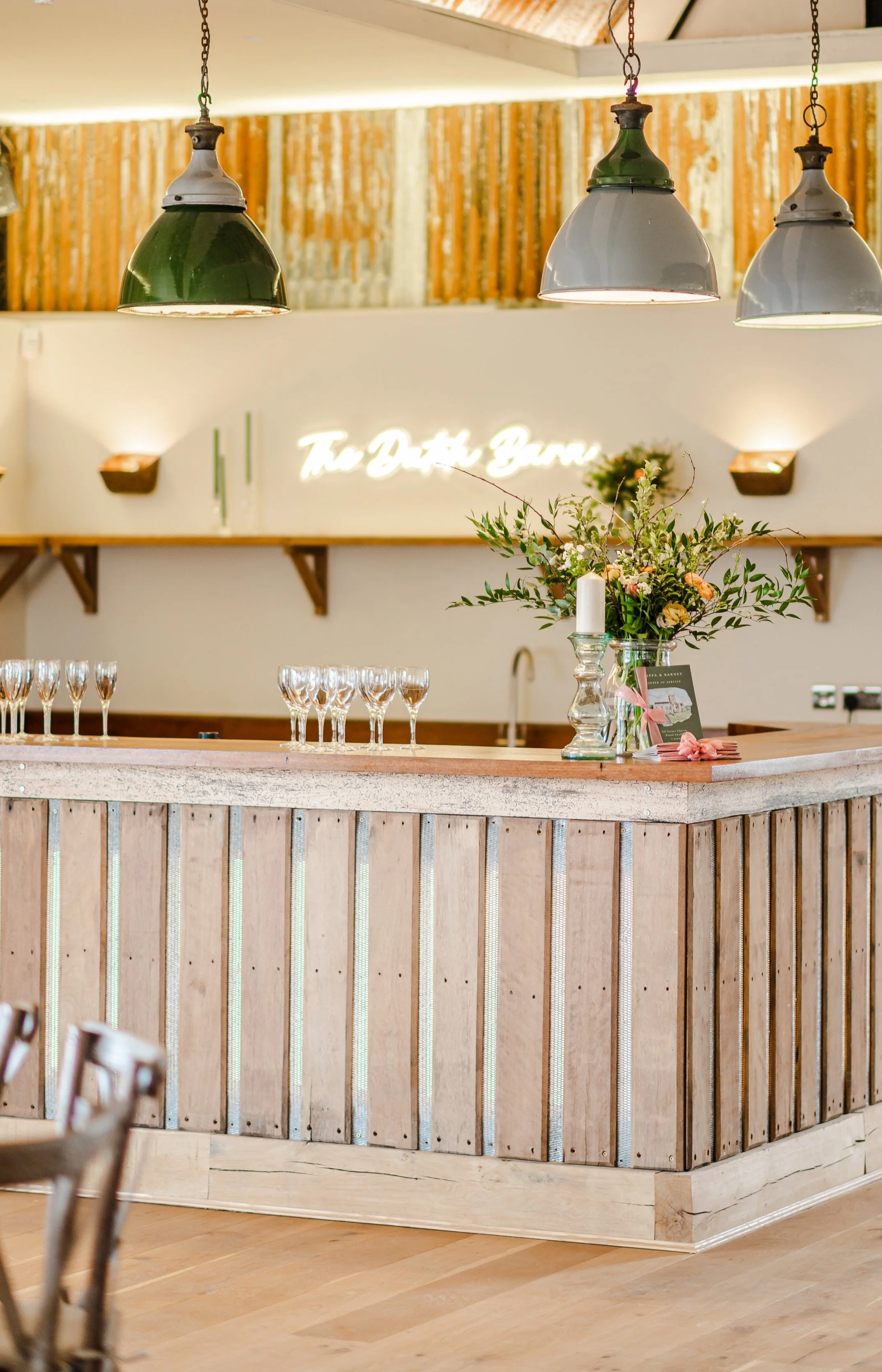 Interior of a bar with a wooden counter, a floral arrangement in a glass vase, wine glasses lined up, and a neon sign in the background that reads 'The Dutch Barn.'