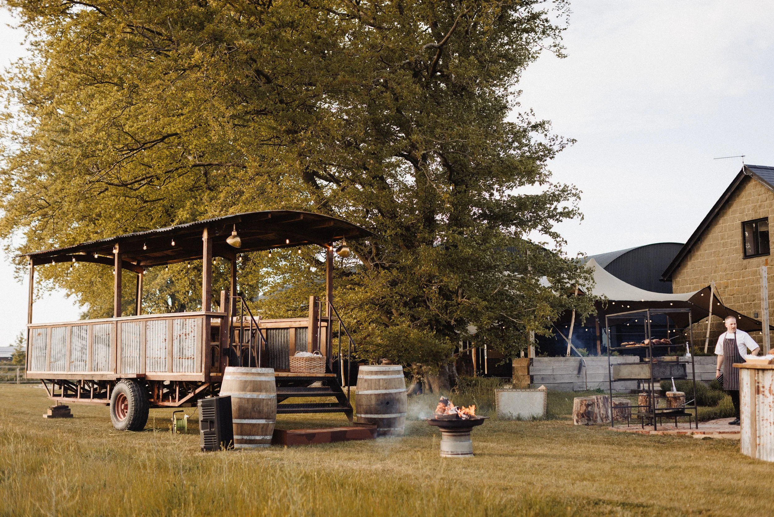 Outdoor dining setup with a wooden stage, barrels, and string lights under a large leafy tree. A person is cooking on a grill with a fire pit nearby.