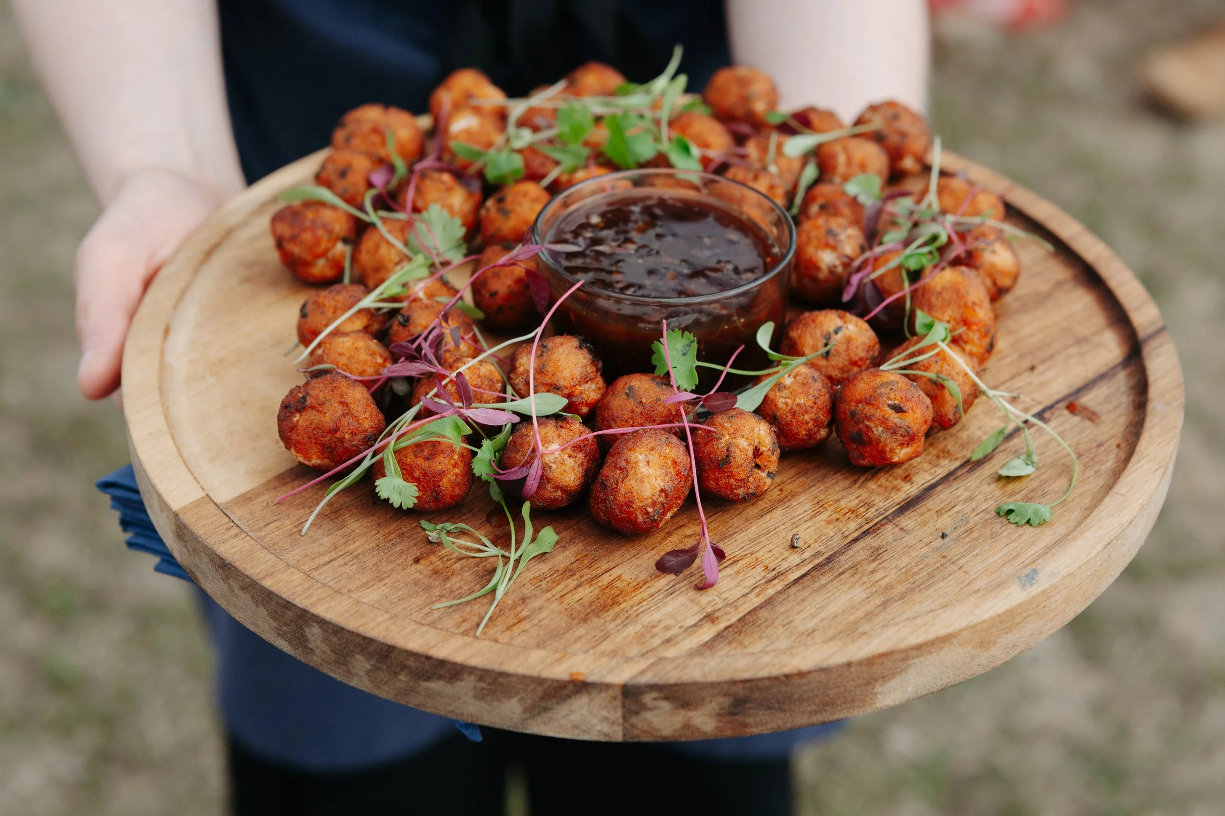 Person holding a wooden platter of fried ball-shaped food garnished with microgreens and a small bowl of dark sauce in the center.