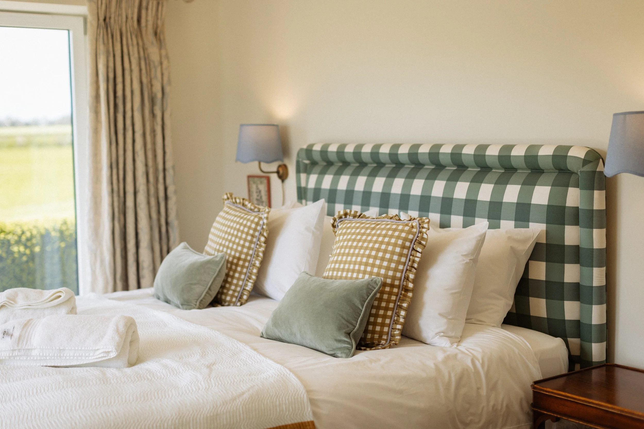 A bedroom with a bed that has a green and white checkered headboard, decorated with plaid pillows and smaller solid-colored pillows. There is a window with patterned curtains and a nightstand with a lamp and a picture frame.