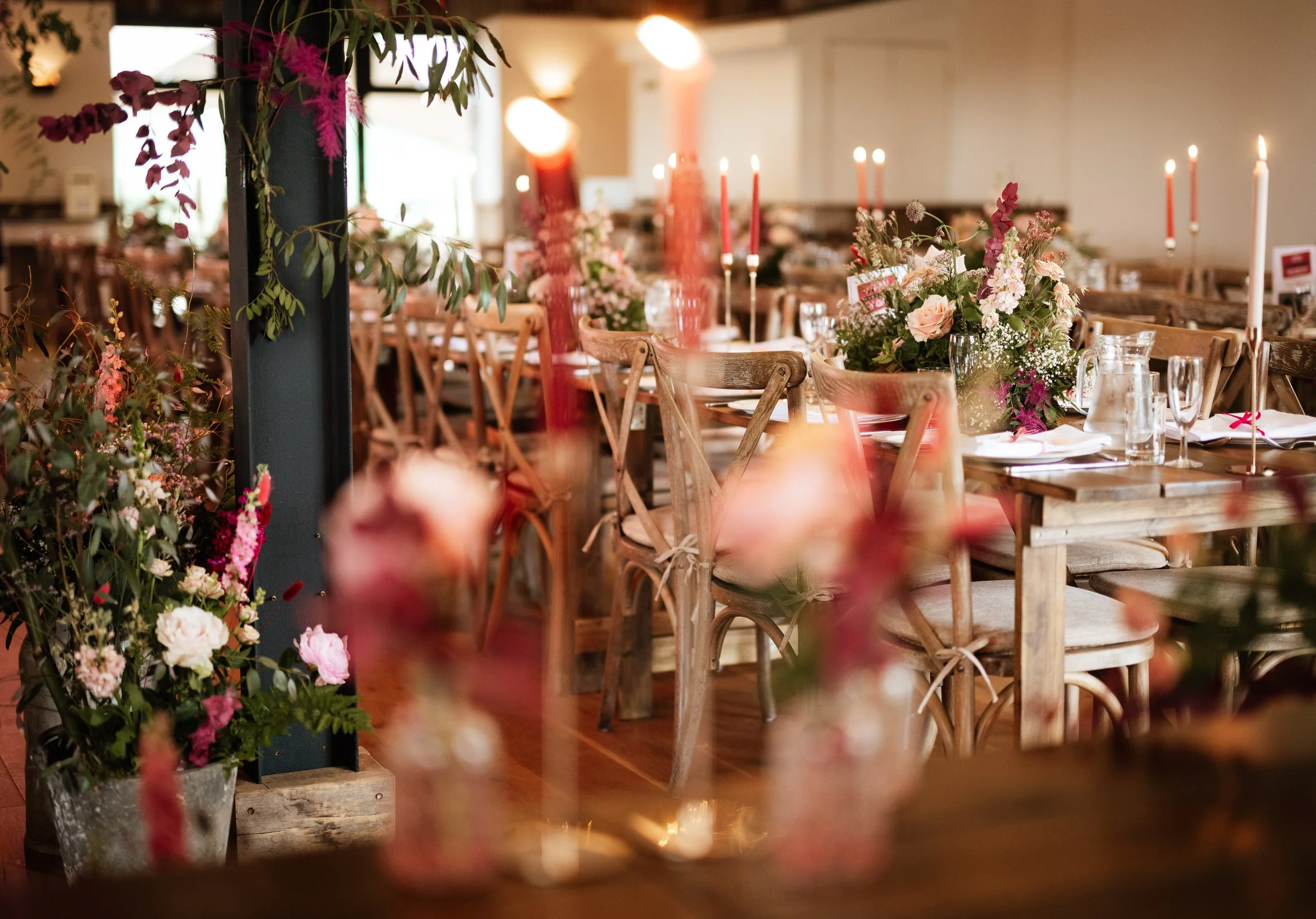 An indoor dining setup with wooden chairs and tables decorated with pink and white floral centerpieces. There are tall pink candles in holders on the tables, and a lush green plant with pink flowers in the foreground. Soft lighting adds a warm ambiance to the scene.