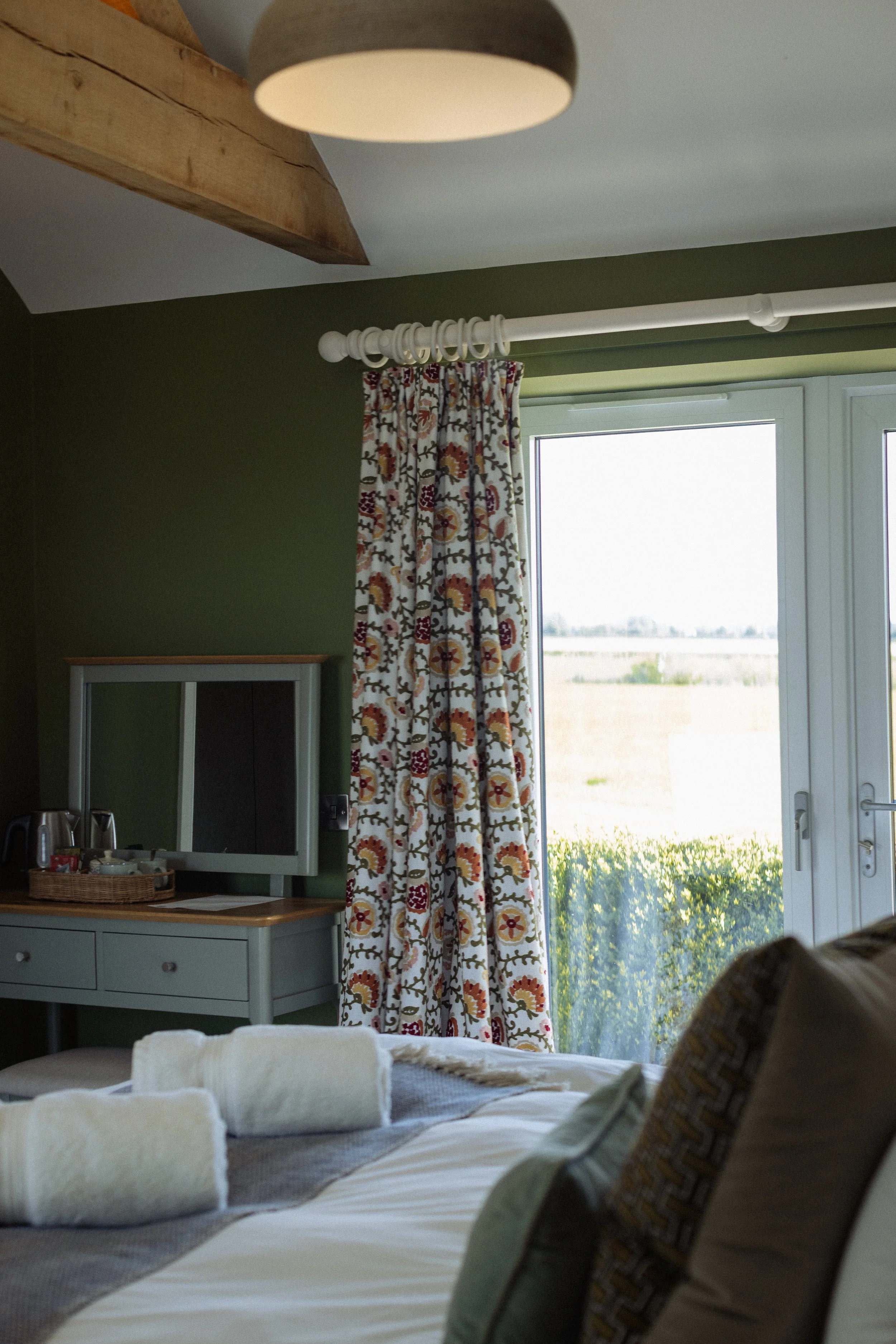 A cozy bedroom with green walls, a large window with floral curtains overlooking a landscape, a white dresser with a mirror, and a bed with neatly folded towels and pillows. There is a ceiling light and wooden beams on the ceiling.
