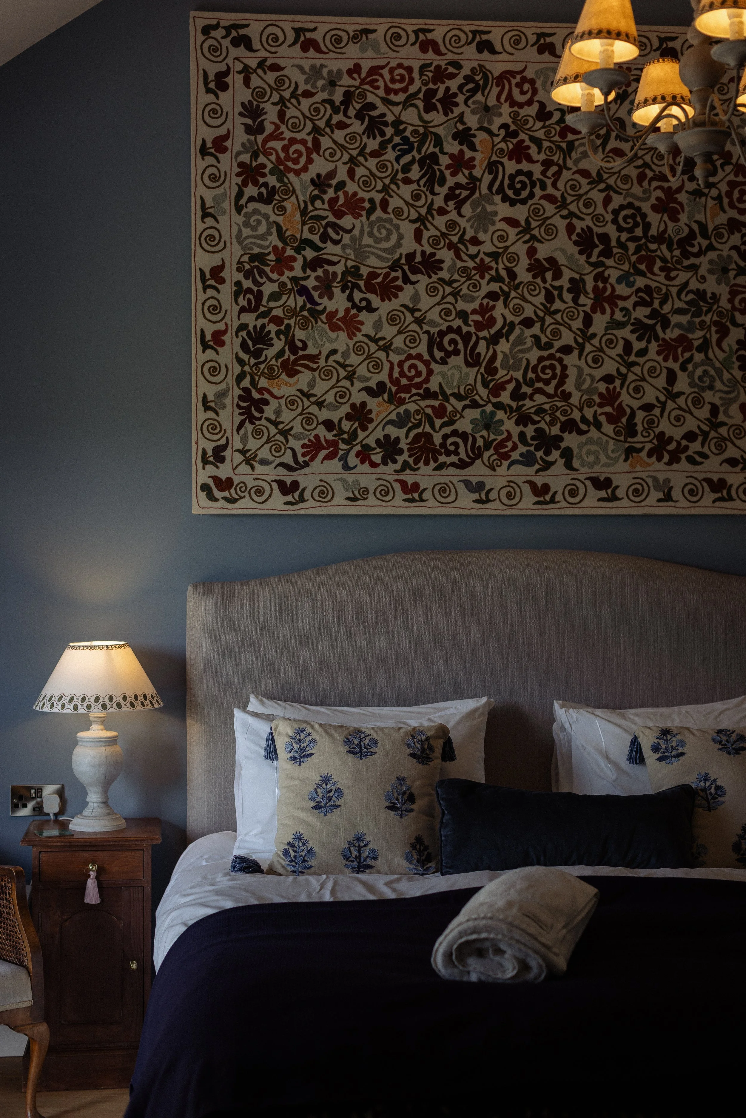 A bedroom with a bed that has white linens and decorative pillows, a wooden nightstand with a white lamp, a rolled towel on the bed, a tapestry with a floral pattern on the wall, and a chandelier with multiple small yellow lamps. The walls are painted a muted blue.