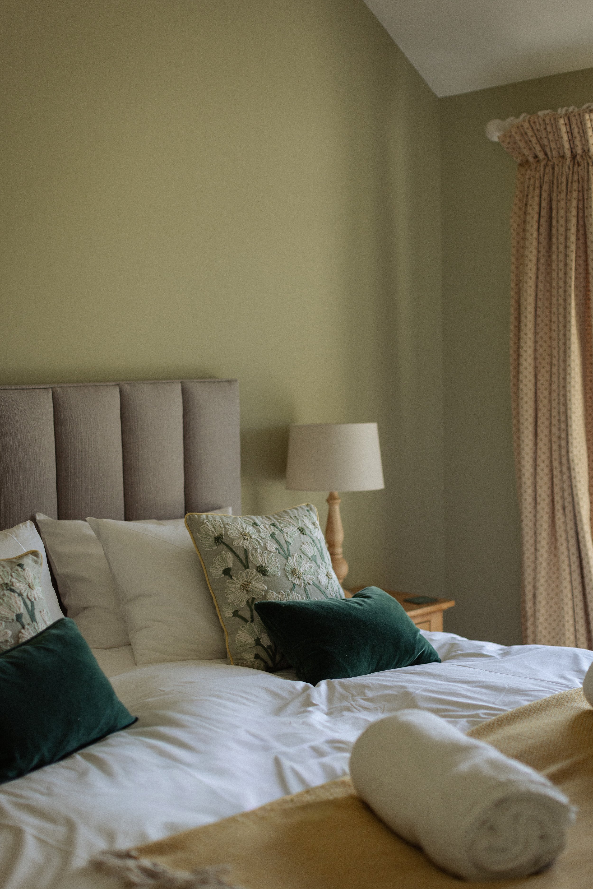 A neatly made bed with white sheets, multiple embroidered and velvet pillows, a beige blanket, and a pillow with a floral design. A rolled-up towel and a white hat are placed at the foot of the bed. There is a beige headboard, a nightstand, and a table lamp next to the bed. The room has light green walls, a wall-mounted light fixture, and curtains with a beige and pink pattern.