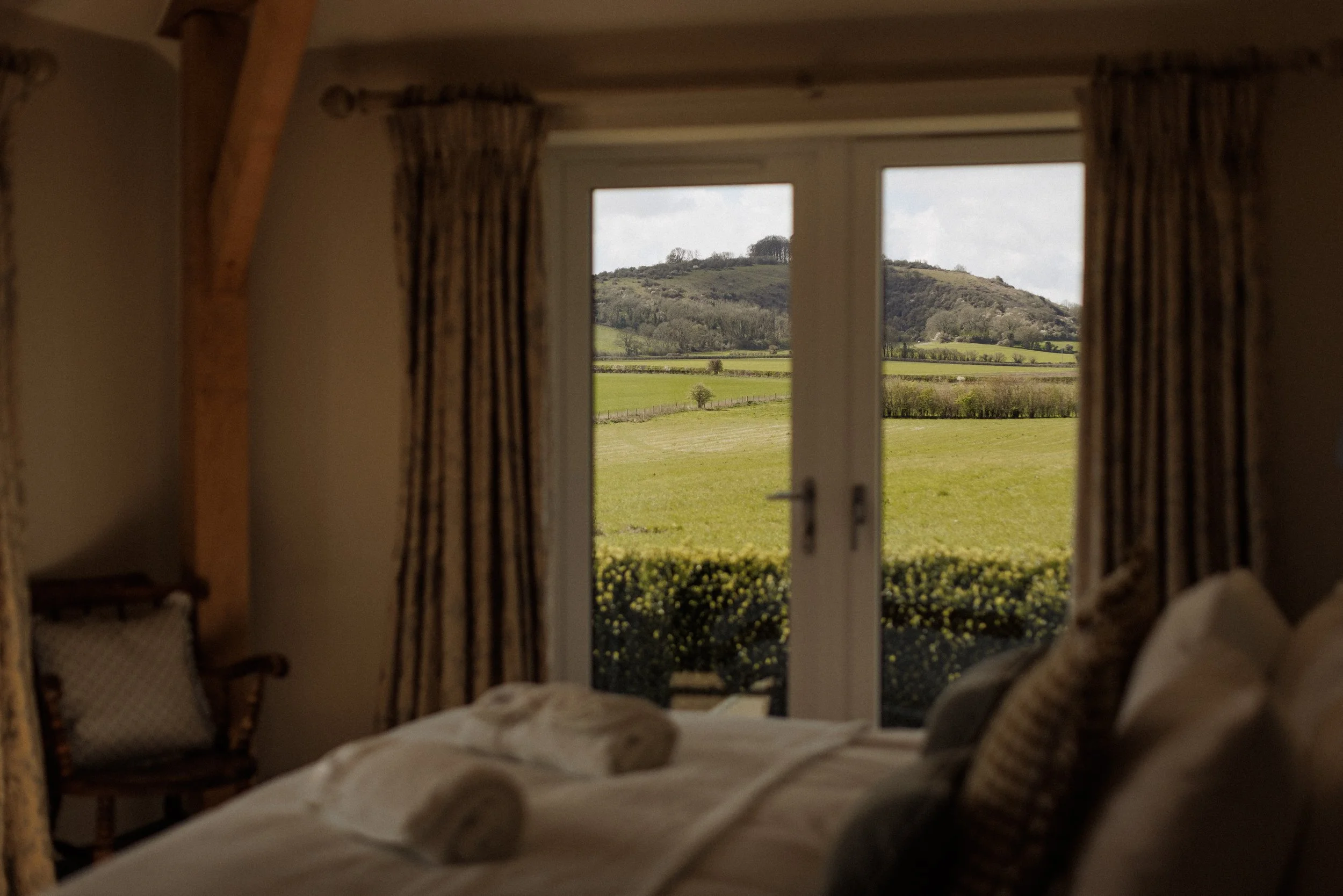 View from inside a house looking out a window at a hillside landscape with trees and fields.