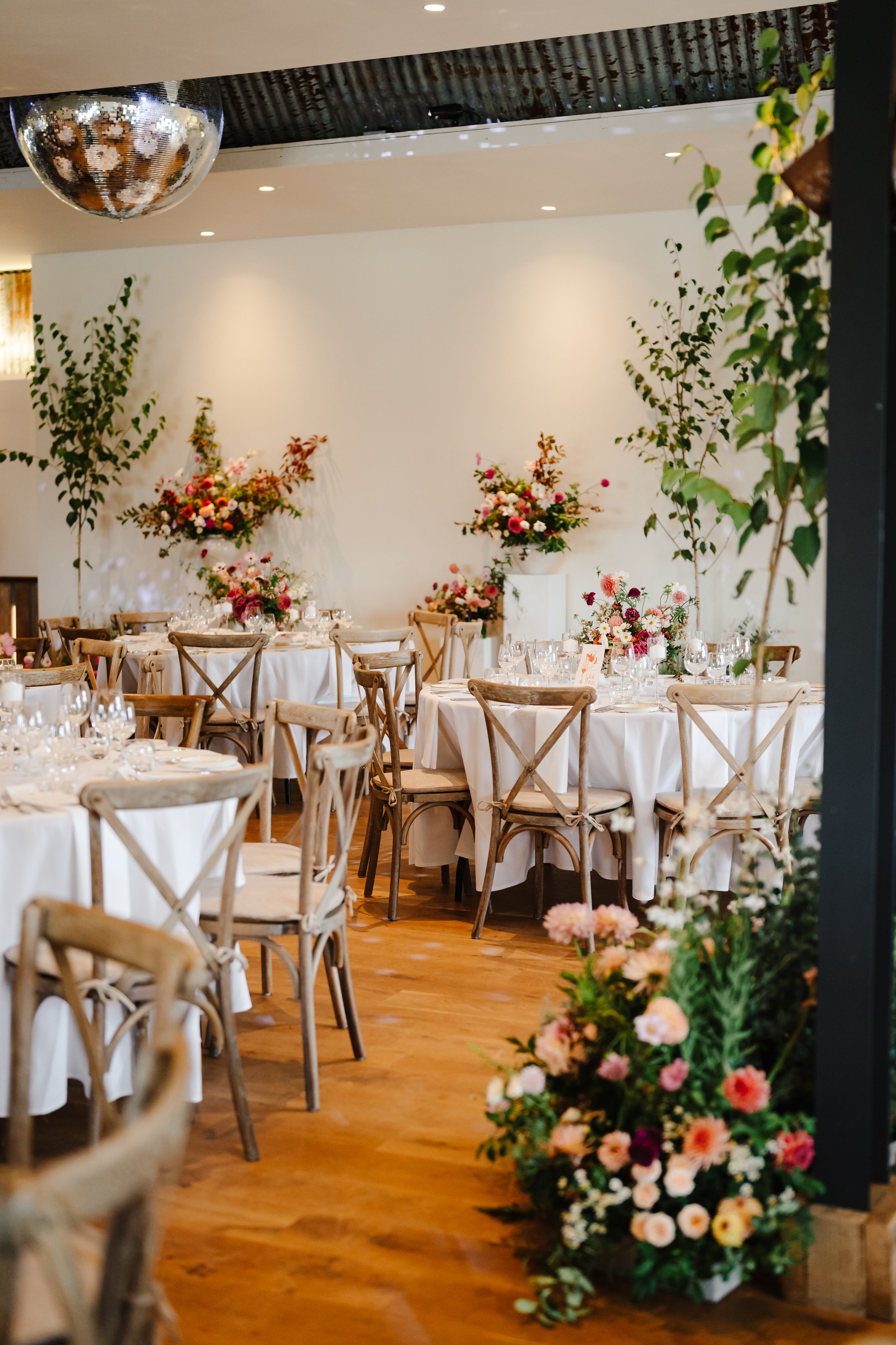 A decorated banquet hall with round tables covered in white tablecloths, set with glassware. The room features large floral arrangements in pink, white, and purple on white pedestals, with green leafy plants around. The floor is wooden, and the lighting is warm, creating an elegant atmosphere for an event.