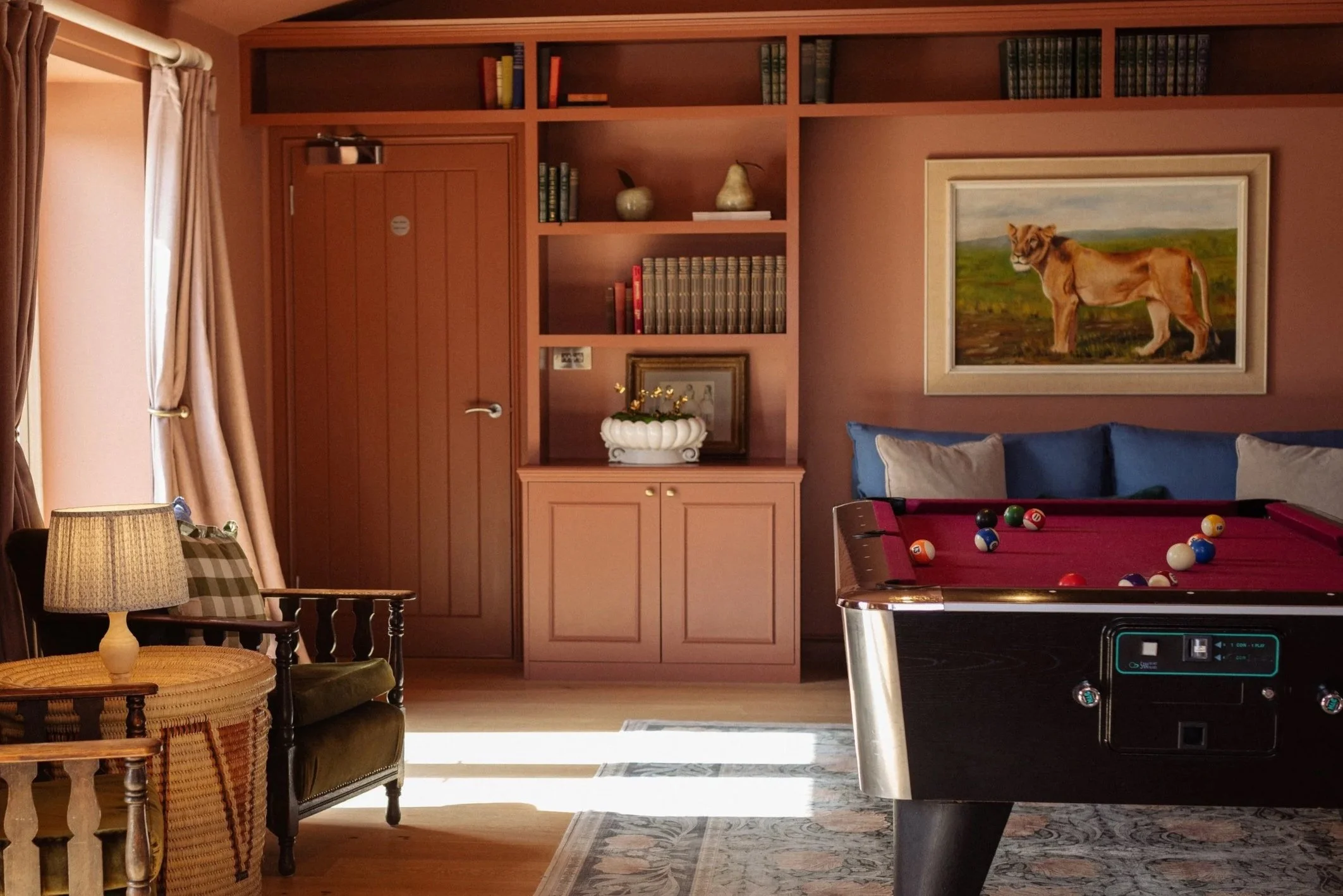 A room with a billiards table and a wall of built-in shelves with books and decorative items, including a framed painting of a lioness.