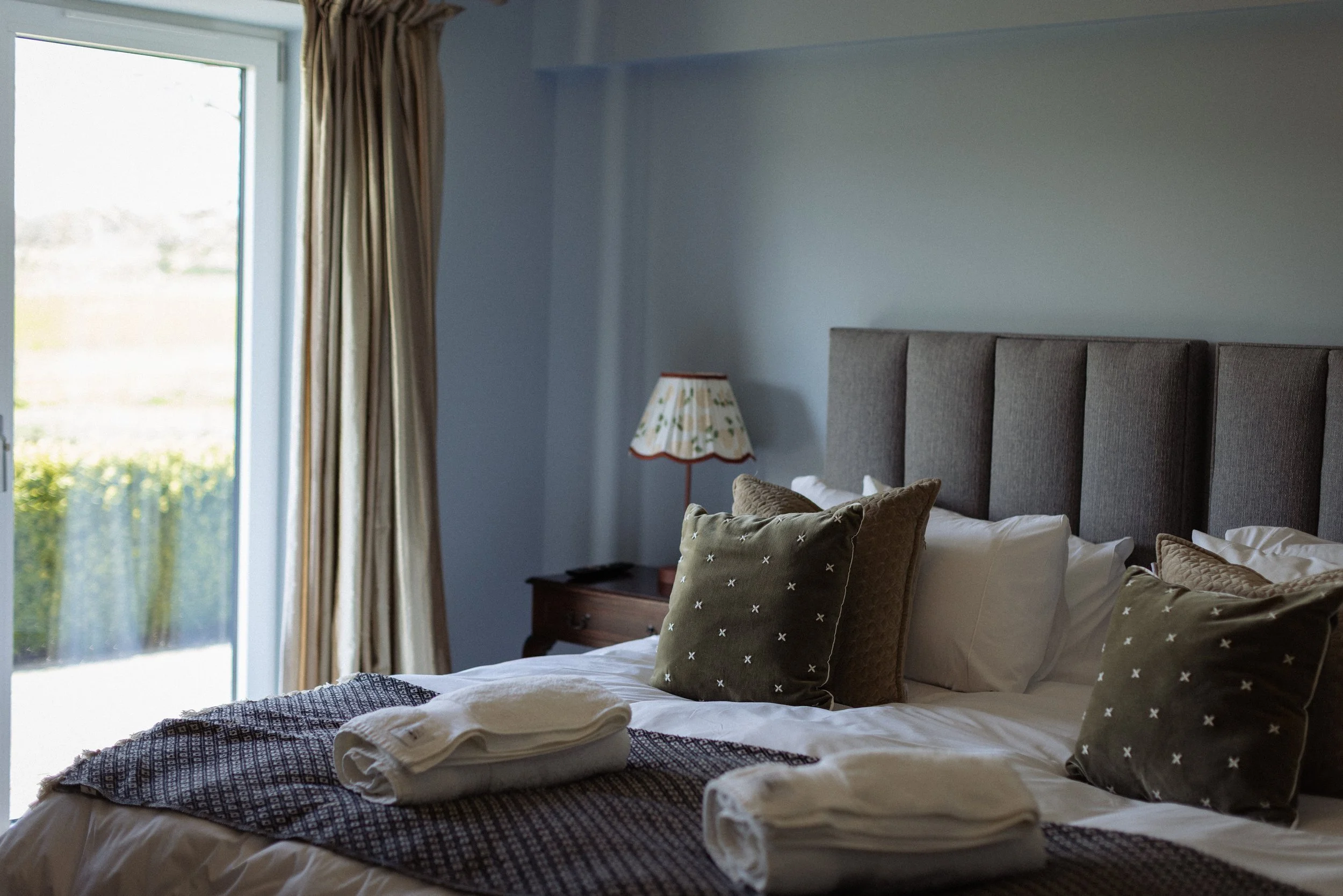 A neatly made bed with beige pillows and folded towels, near a window with curtains and a bedside lamp in a cozy bedroom.