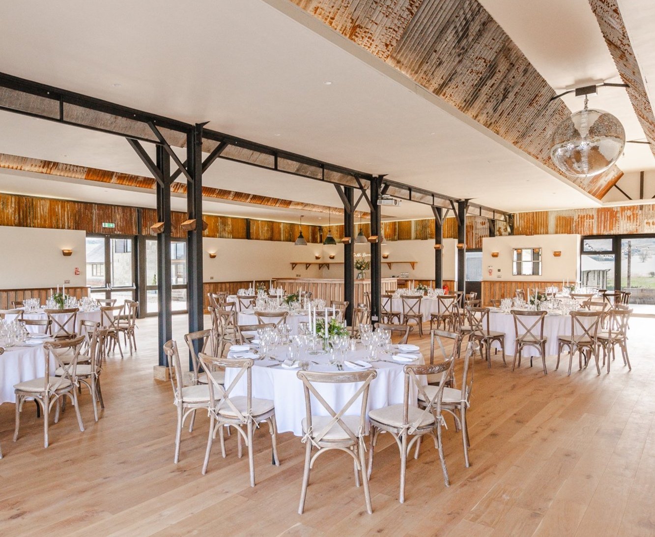Well-decorated banquet hall with round tables covered in white tablecloths, set with glassware, silverware, and floral centerpieces, featuring wooden chairs and large windows letting in natural light.
