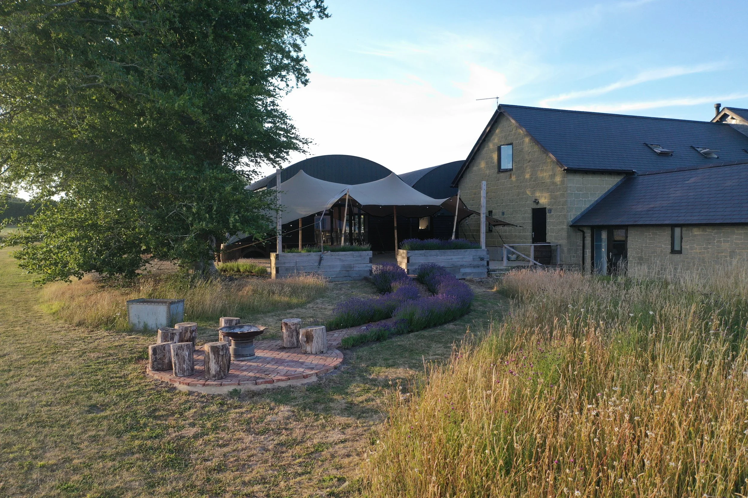 A rural scene featuring a stone house with a sloped roof, a large tree on the left, a white canopy tent in the center, and a garden with purple flowers and a circular brick seating area in the foreground under a clear blue sky.