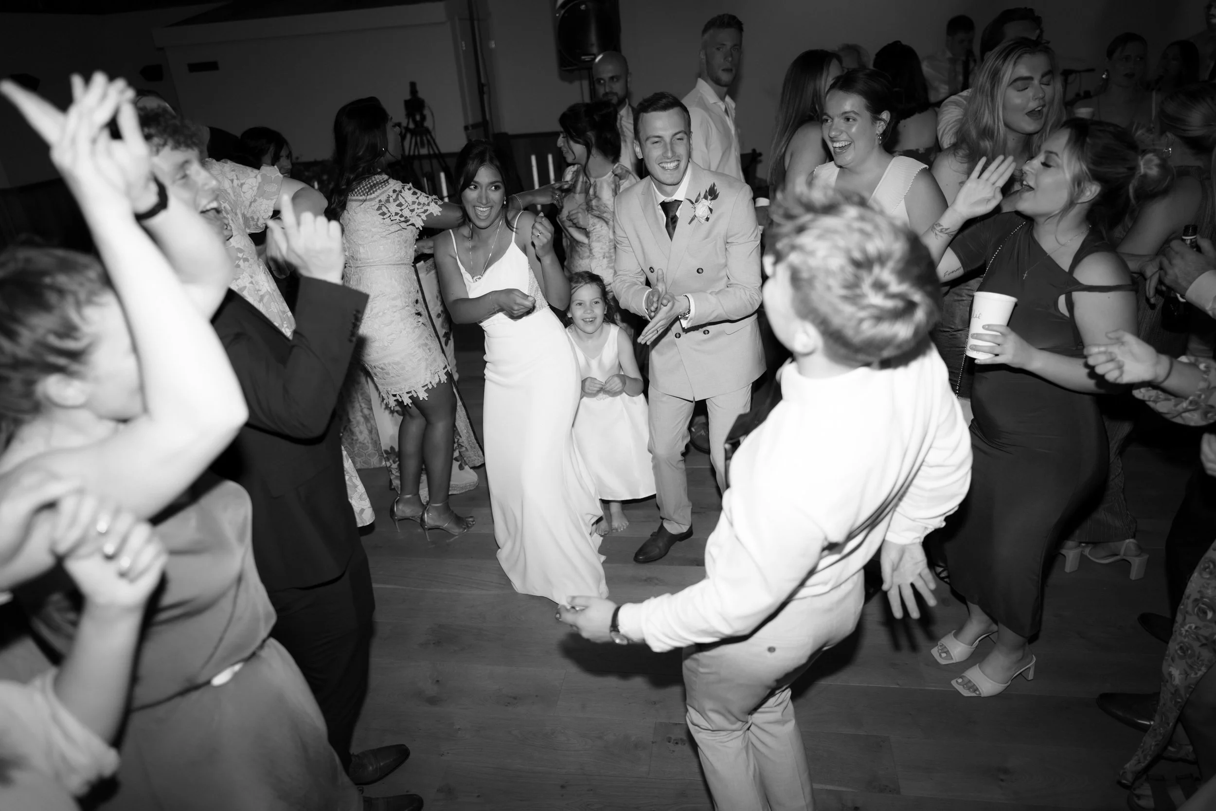People dancing and celebrating at a wedding reception, including a woman in a white dress, a man in a suit with a boutonniere, and various guests smiling and enjoying the event.