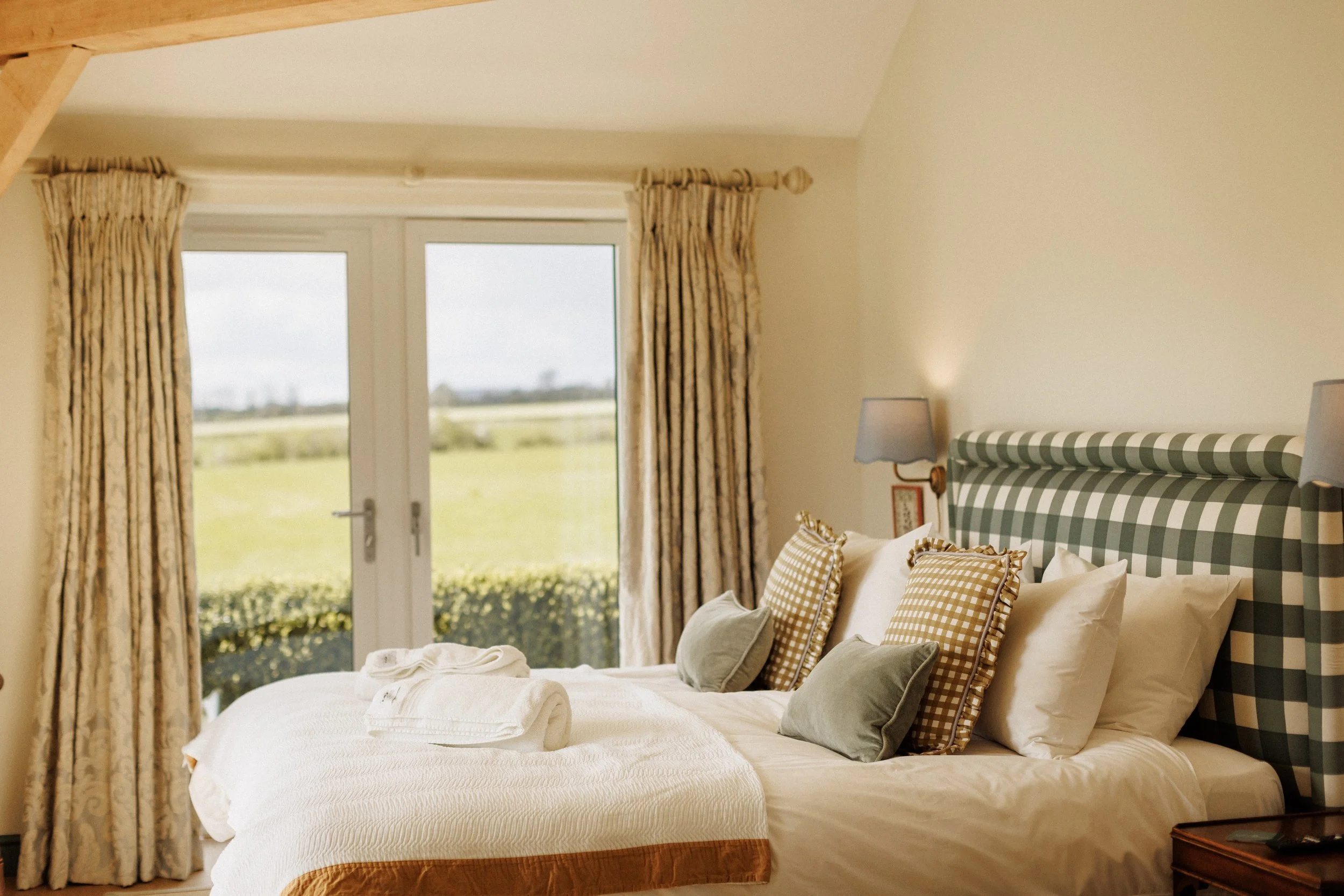 A neatly made bed with white pillows and green velvet cushions in a cozy room with a window and curtains, beige walls, and a wall-mounted lamp.