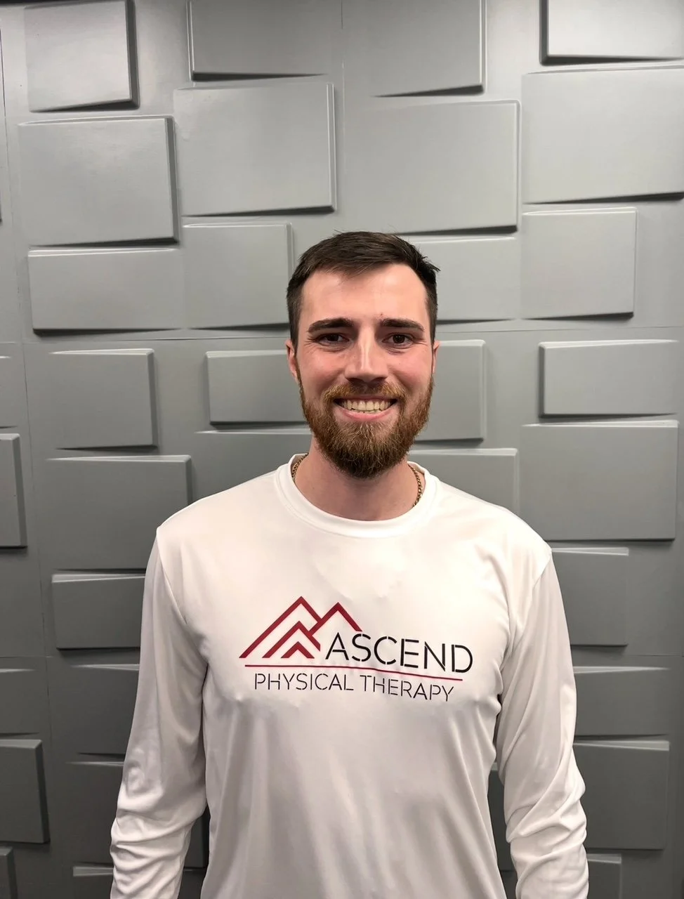 A young man with a beard smiling at the camera, wearing a white long-sleeve shirt with the logo and name of 'Ascend Physical Therapy' printed on it, standing in front of a gray textured wall.