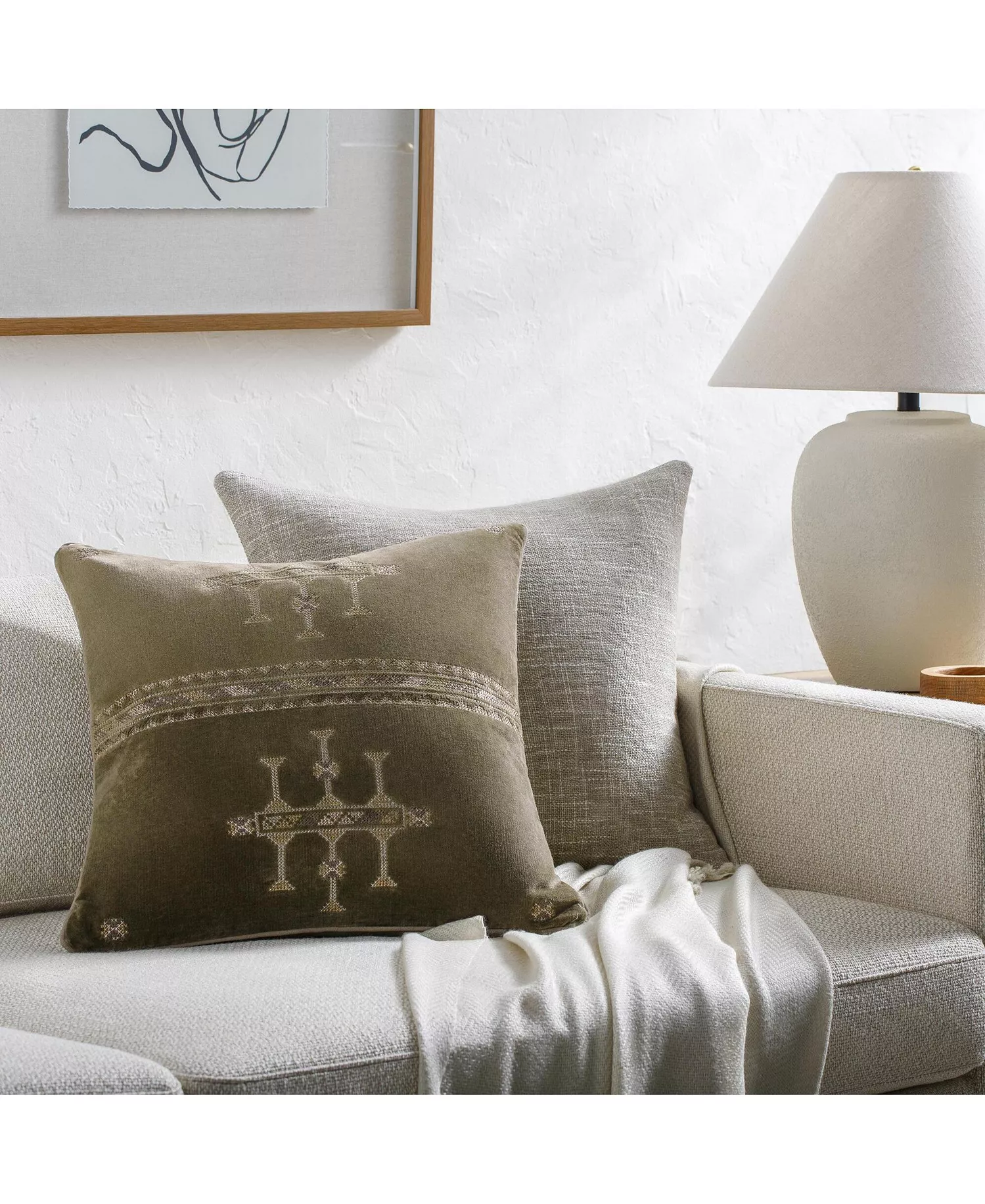 Velour Decorative Pillow by Surya, available through Bloomingdale’s, featuring a muted olive green finish and delicate geometric embroidery in warm neutral tones. Its subtle sheen and earthy palette add depth to cream upholstery, bringing an inviting