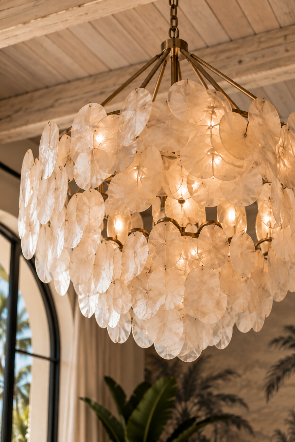 Elegant chandelier made of translucent, round, shell-like discs with warm lighting, hanging from a chain in a room with large arched window and tropical plants.