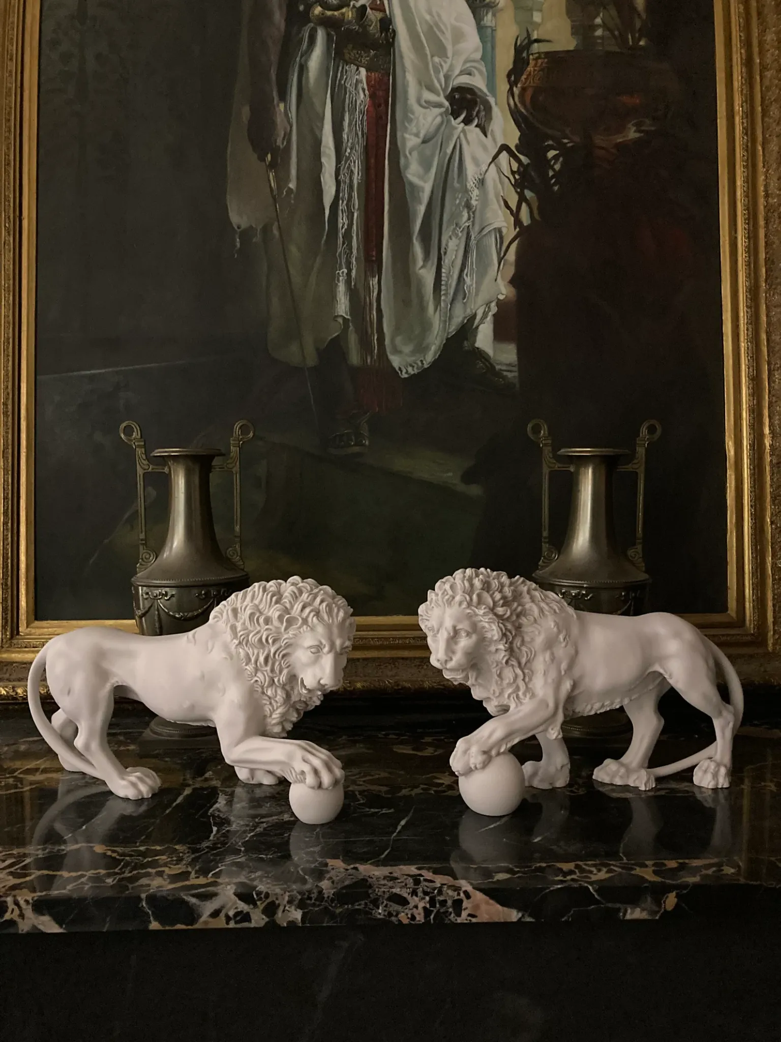 Pair of white Medici and Vacca lion sculptures with paws resting on spheres, displayed on dark marble beneath a gilt-framed painting.