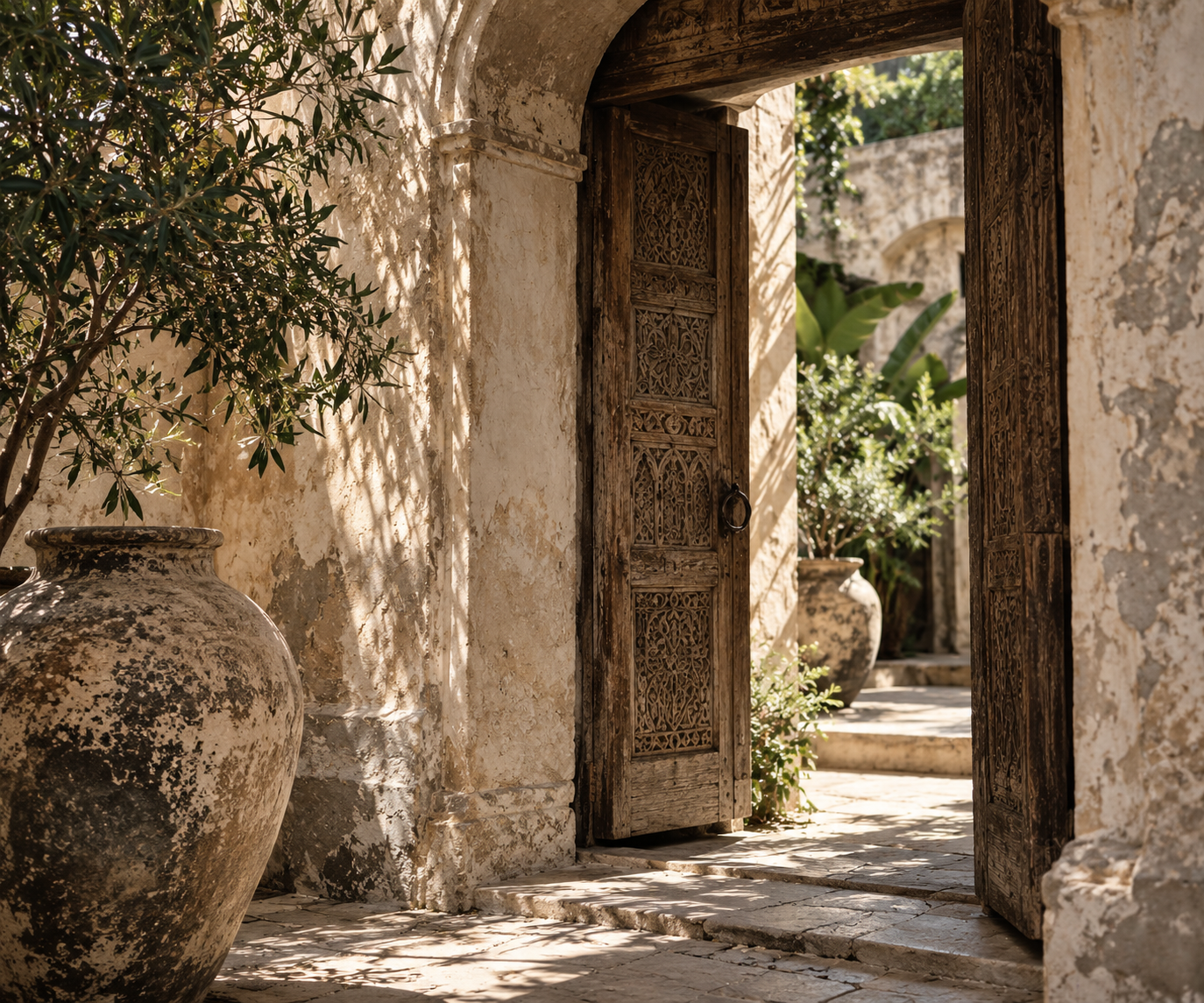 Weathered carved wood doorway framed by aged plaster, stone and greenery in a sunlit old-world courtyard.