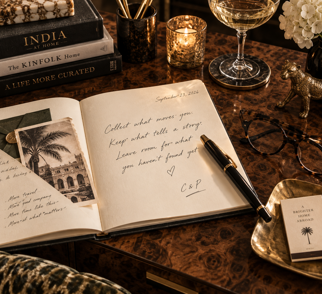 A desk with a journal open and a handwritten note, books, candles, a glass of white wine, a pair of glasses, a small decorative leopard sculpture, photographs, and a gold tray with a card that reads 'A Brighter Home Abroad'.