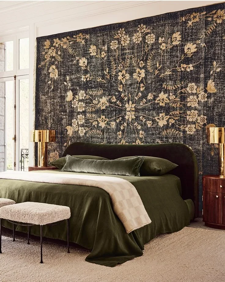 A bedroom with a dark green bed, matching pillows, and a beige throw blanket. Behind the bed, a large blue and white floral-patterned rug hangs as a wall tapestry. Two brass bedside lamps and a small wooden side table are also visible.