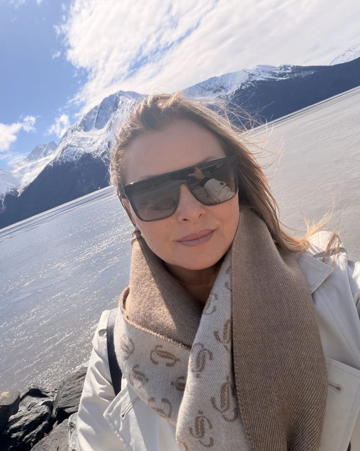 A woman taking a selfie outdoors with snow-covered mountains and a partly cloudy sky in the background.