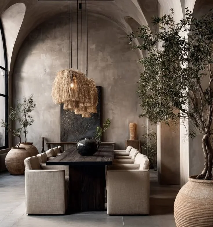 Elegant dining room with a dark wooden table surrounded by cream upholstered chairs, large potted plants, textured wall art, and a woven bamboo pendant light.