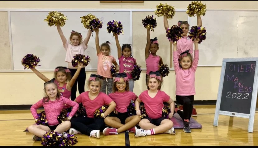Group of young girls cheerleaders in pink and purple outfits holding purple and gold pom-poms in a gymnasium, with a chalkboard reading 'CHEER MADNESS 2022'.