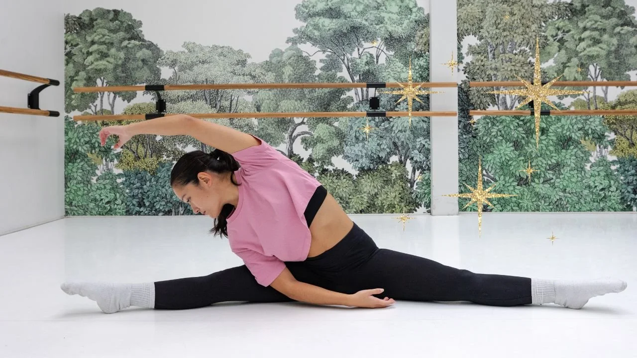 A woman practicing yoga in a studio with a mural of trees on the wall, performing a side stretch stretch in a seated split position. There are wooden ballet barres and decorative star-like effects added to the image.