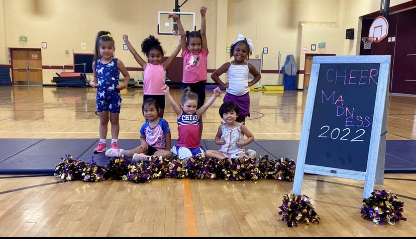 Seven young girls in cheerleading outfits and casual clothing in a gymnasium, posing cheerfully with some standing and some sitting on the floor with pom-poms, a blackboard with colorful writing, and a hardwood basketball court in the background.