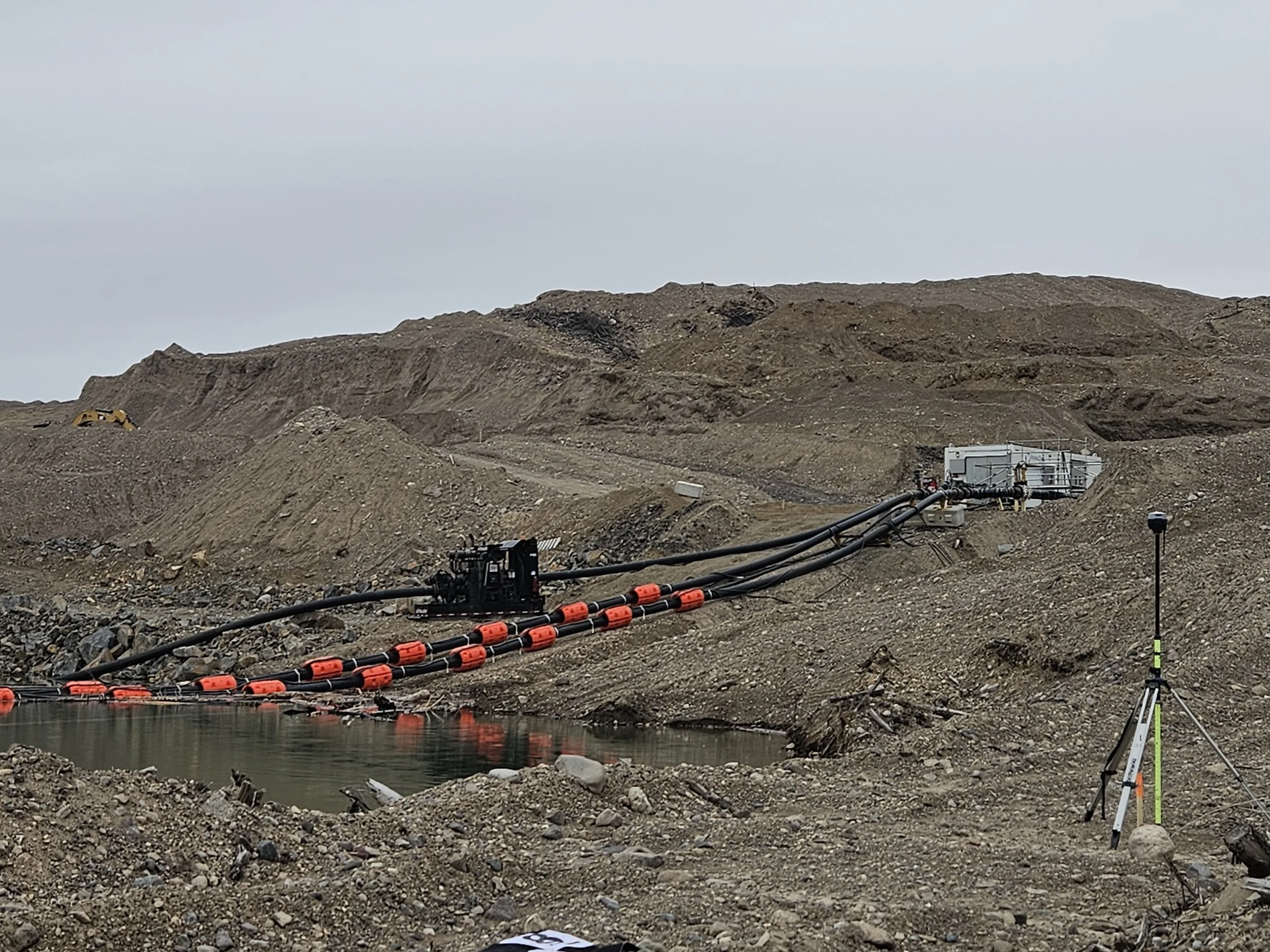 Construction site with excavated dirt hills, a body of water at the foreground, and equipment including pipes, tape barriers, and surveying tools.