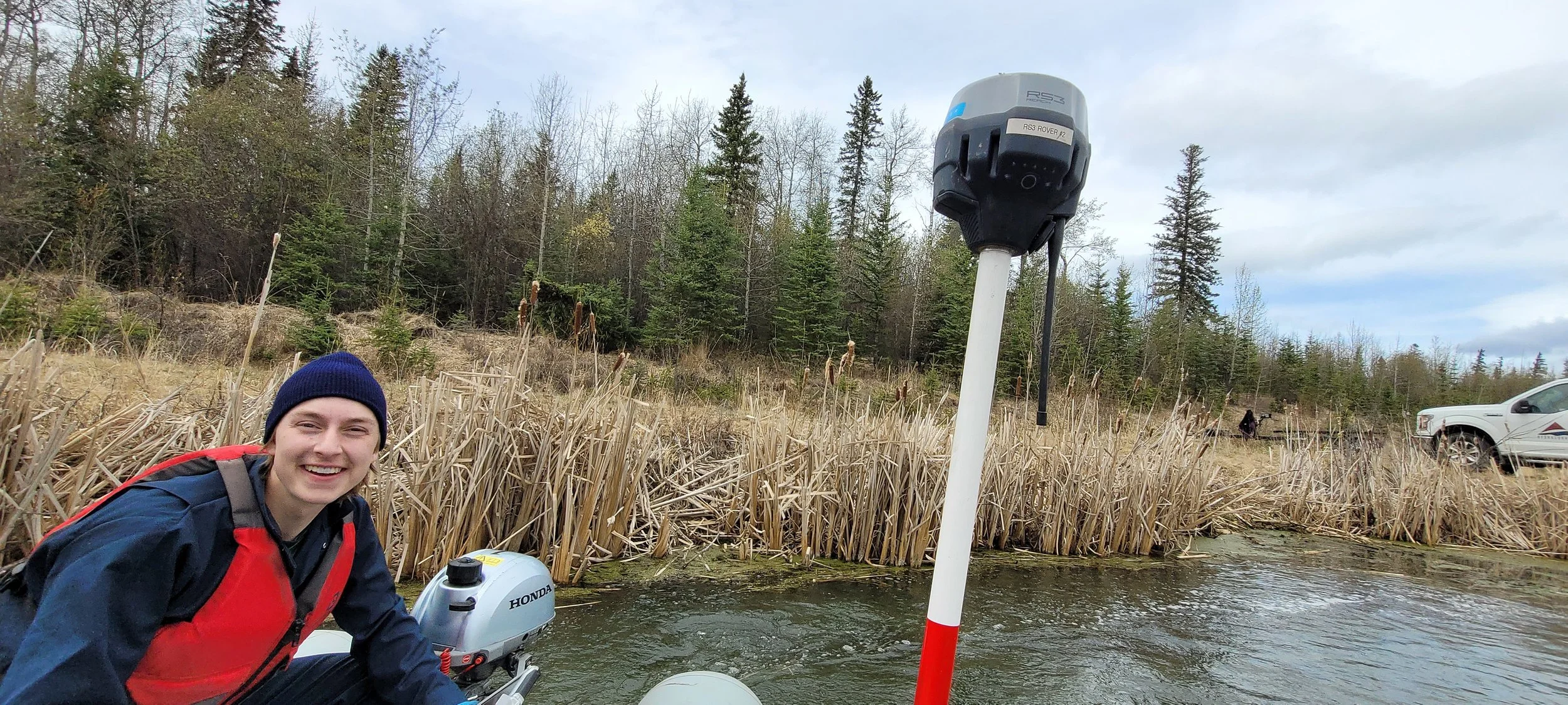 A person wearing a  red life jacket smiling near the water, with a hydrographic survey device mounted on a pole and a Honda outboard motor visible. The background features dry grass, trees, and a partly cloudy sky.