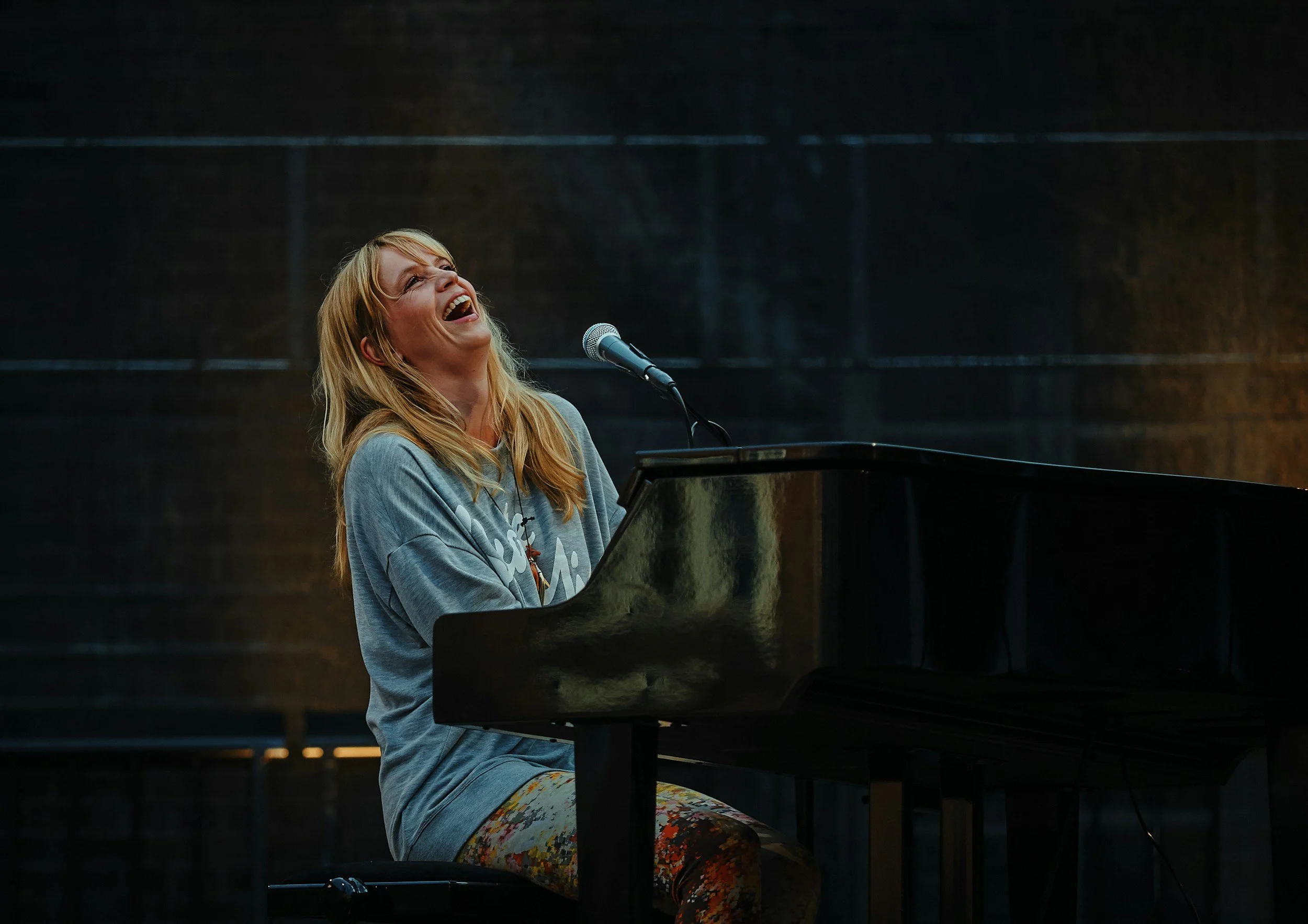 A woman with blonde hair laughing and playing a grand piano with a microphone on a stage.