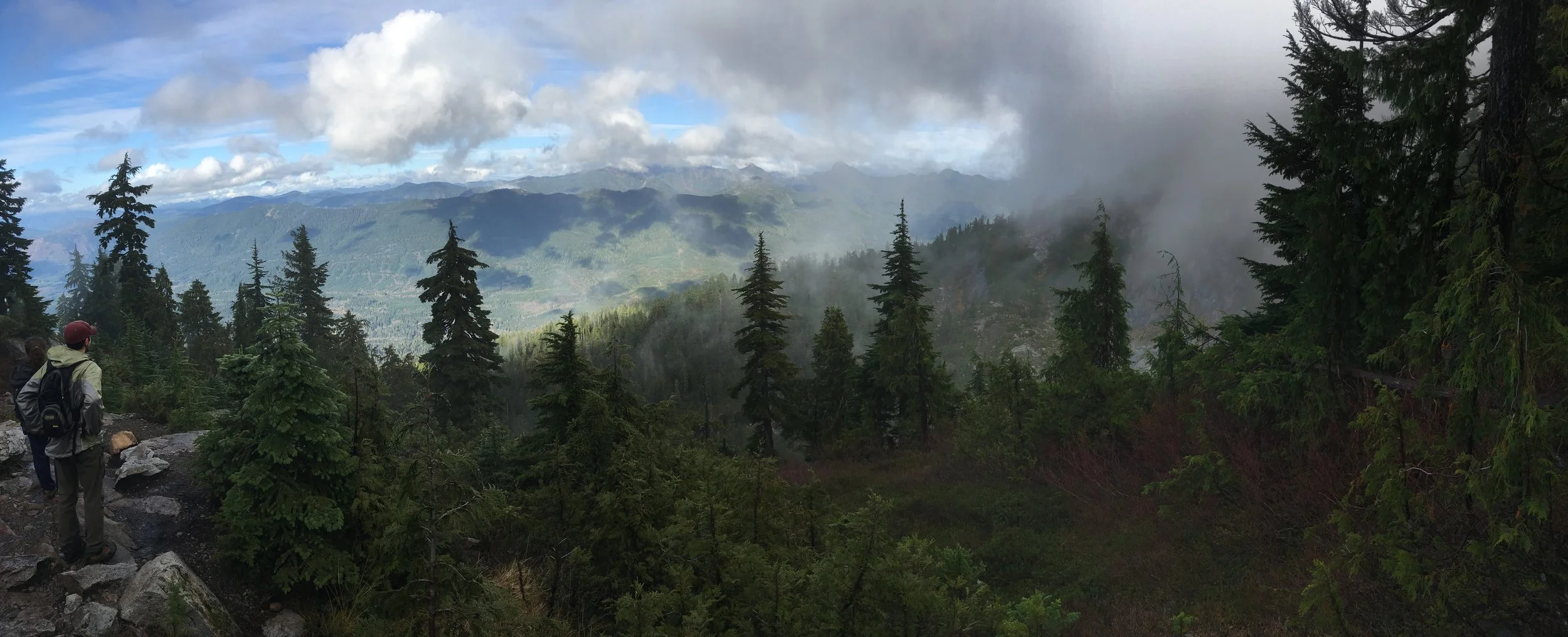 Two hikers standing on a rocky trail in a dense green forest, overlooking mountains with clouds and mist in the background.