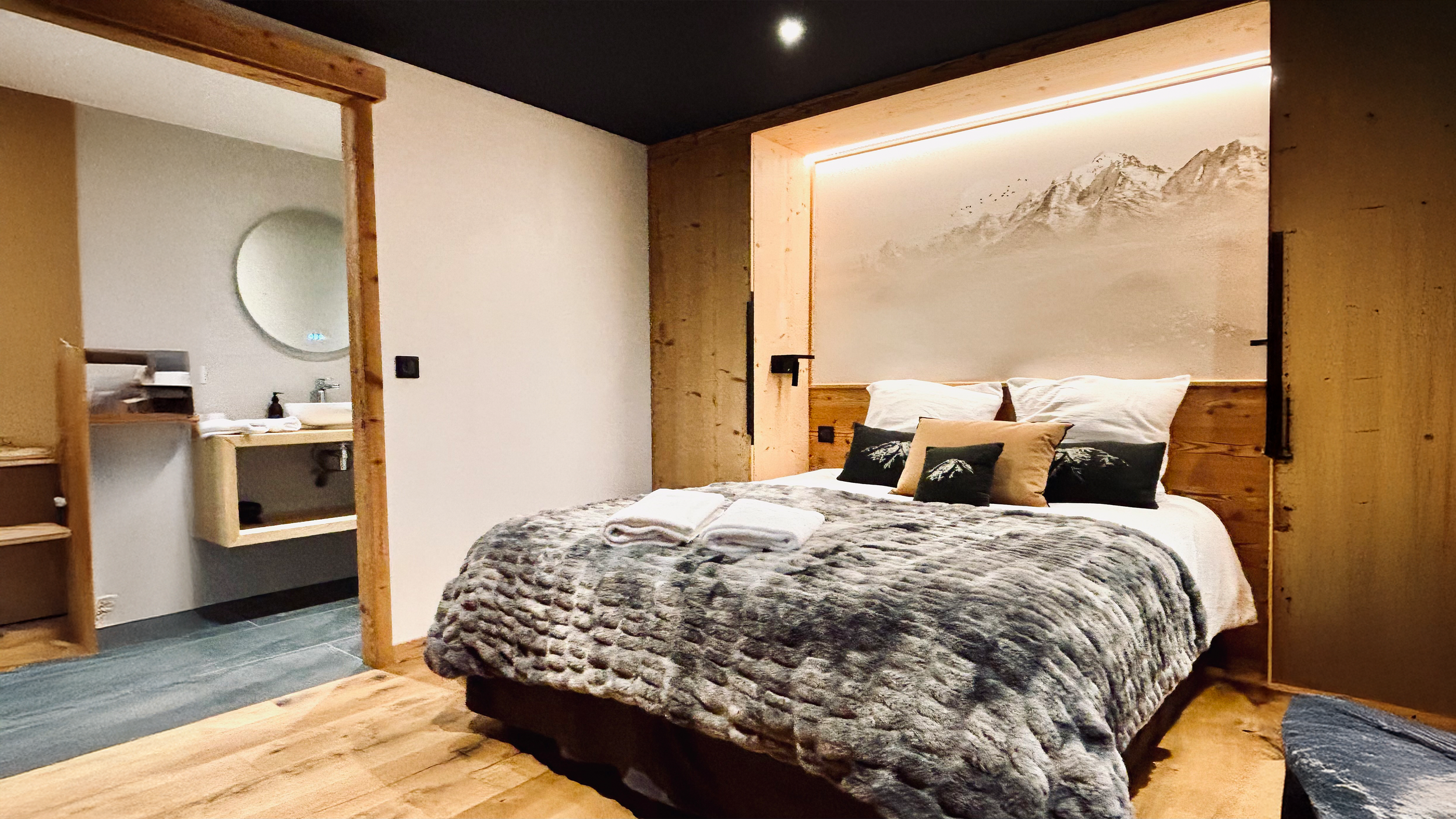 Chambre à coucher avec un lit King size, décor bois clair, mur avec une fresque de montagnes, et une porte coulissante en bois menant à une salle de bain.