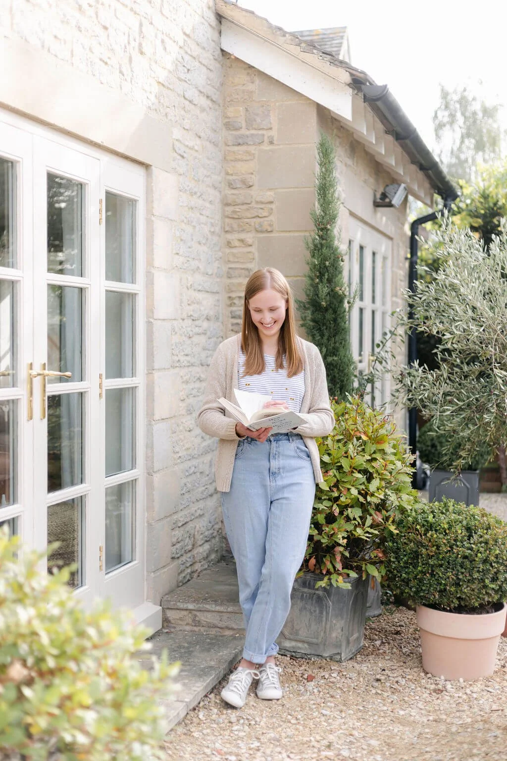 Therapist standing outside a stone house, reading a book, surrounded by potted plants and greenery in a garden setting.