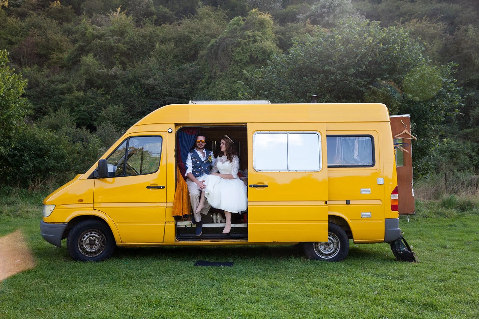 A couple dressed in wedding attire sitting inside a yellow van with the side door open, parked on a grassy field with bushes and trees in the background.