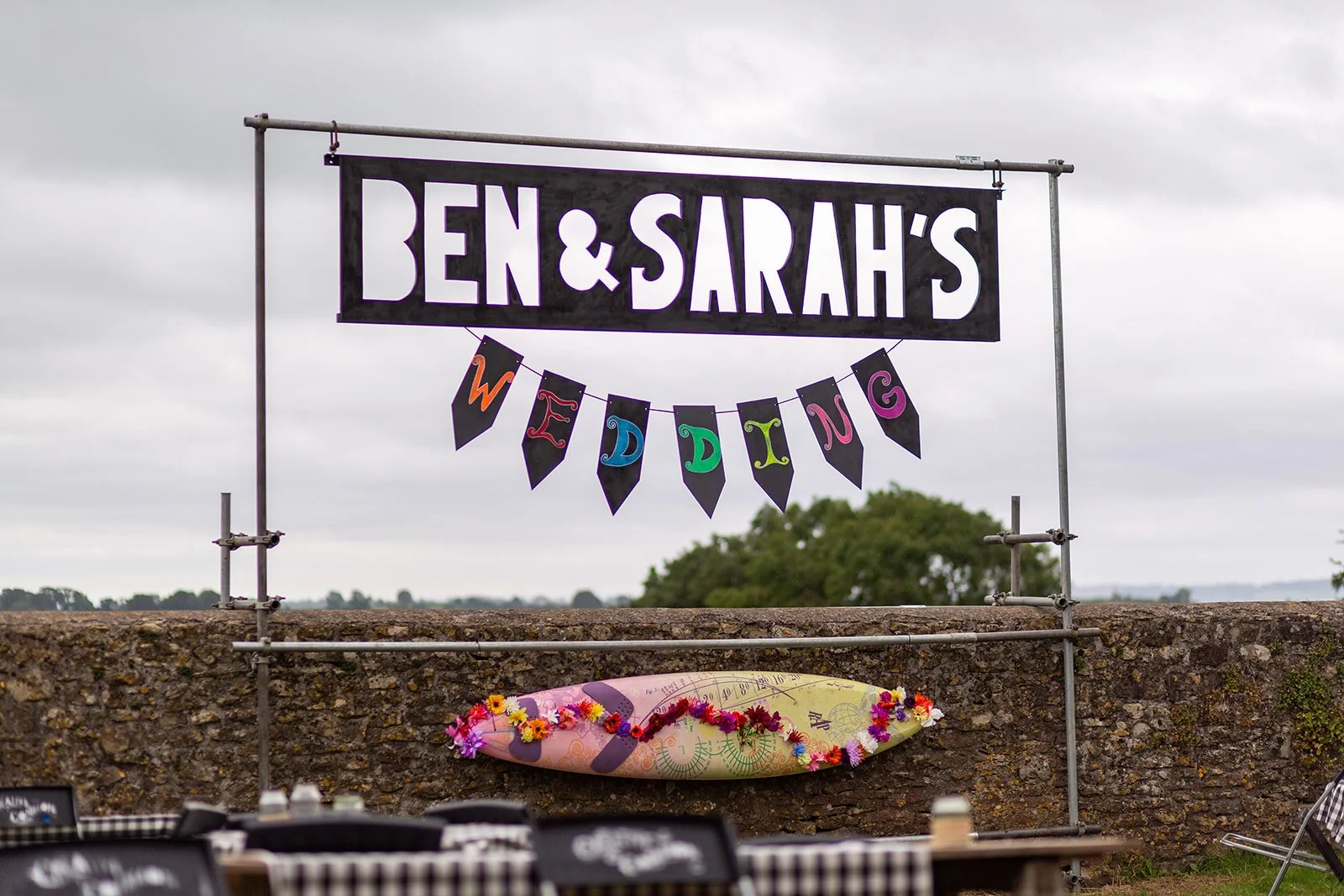 Sign for Ben & Sarah's wedding with colorful 'Wedding' banner and floral surfboard under cloudy sky.
