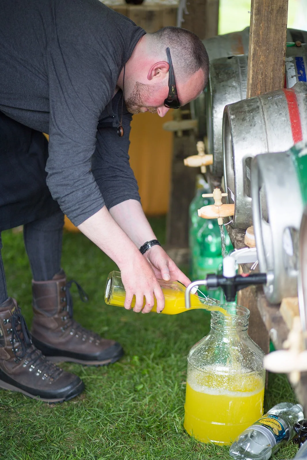 A man pouring orange juice from a glass bottle into a glass jar under an outdoor faucet, with other juice bottles and a water bottle on the grass nearby.