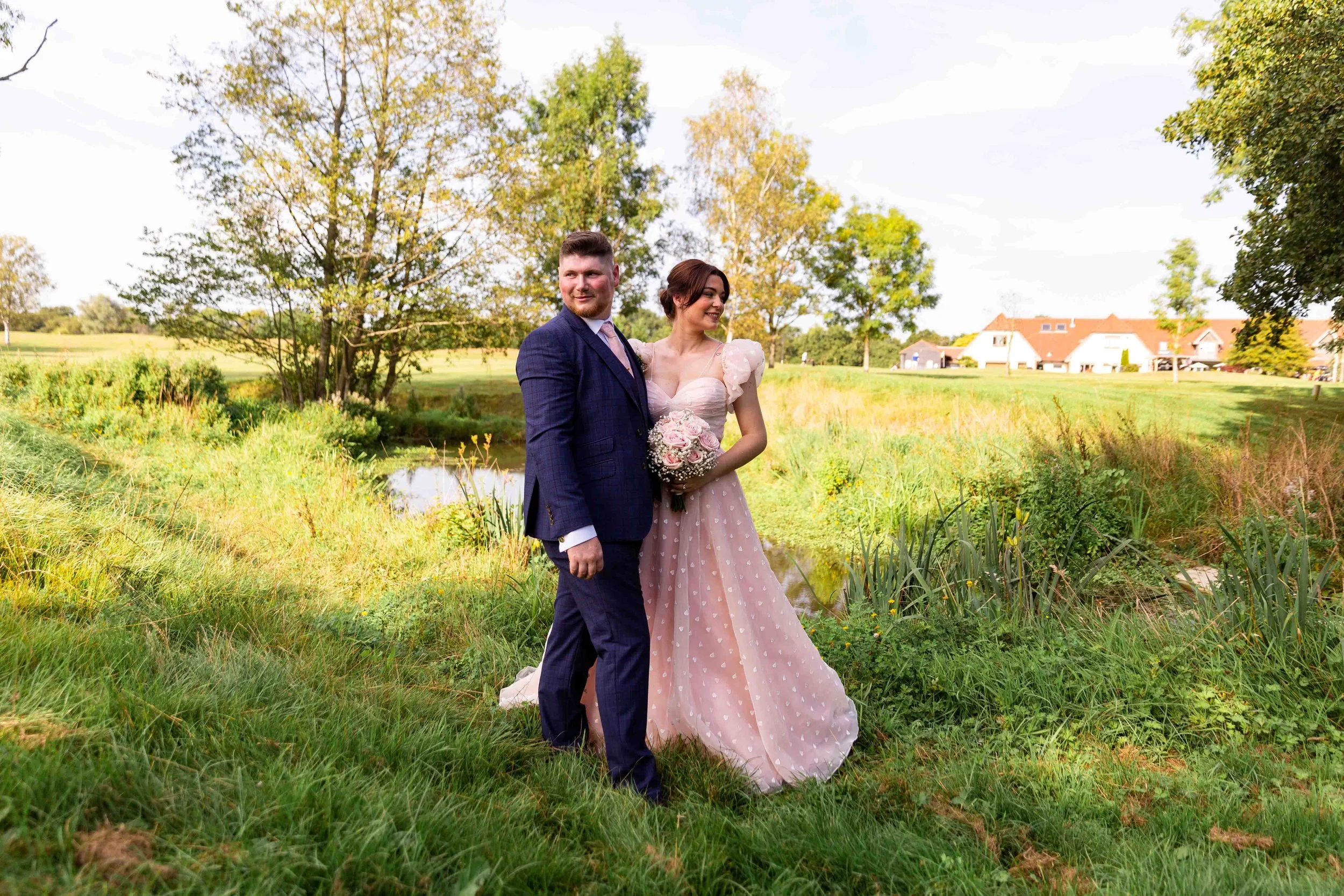 A newlywed couple standing beside a small pond in a green park on a sunny day, with trees and houses in the background.