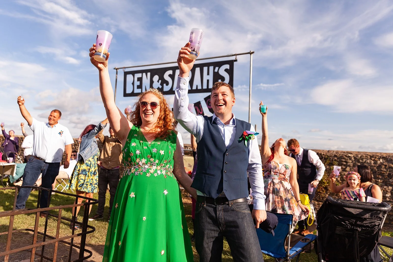People celebrating outdoors at a wedding, holding drinks, with a sign that says 'Ben & Sarah's' in the background, under a partly cloudy sky.