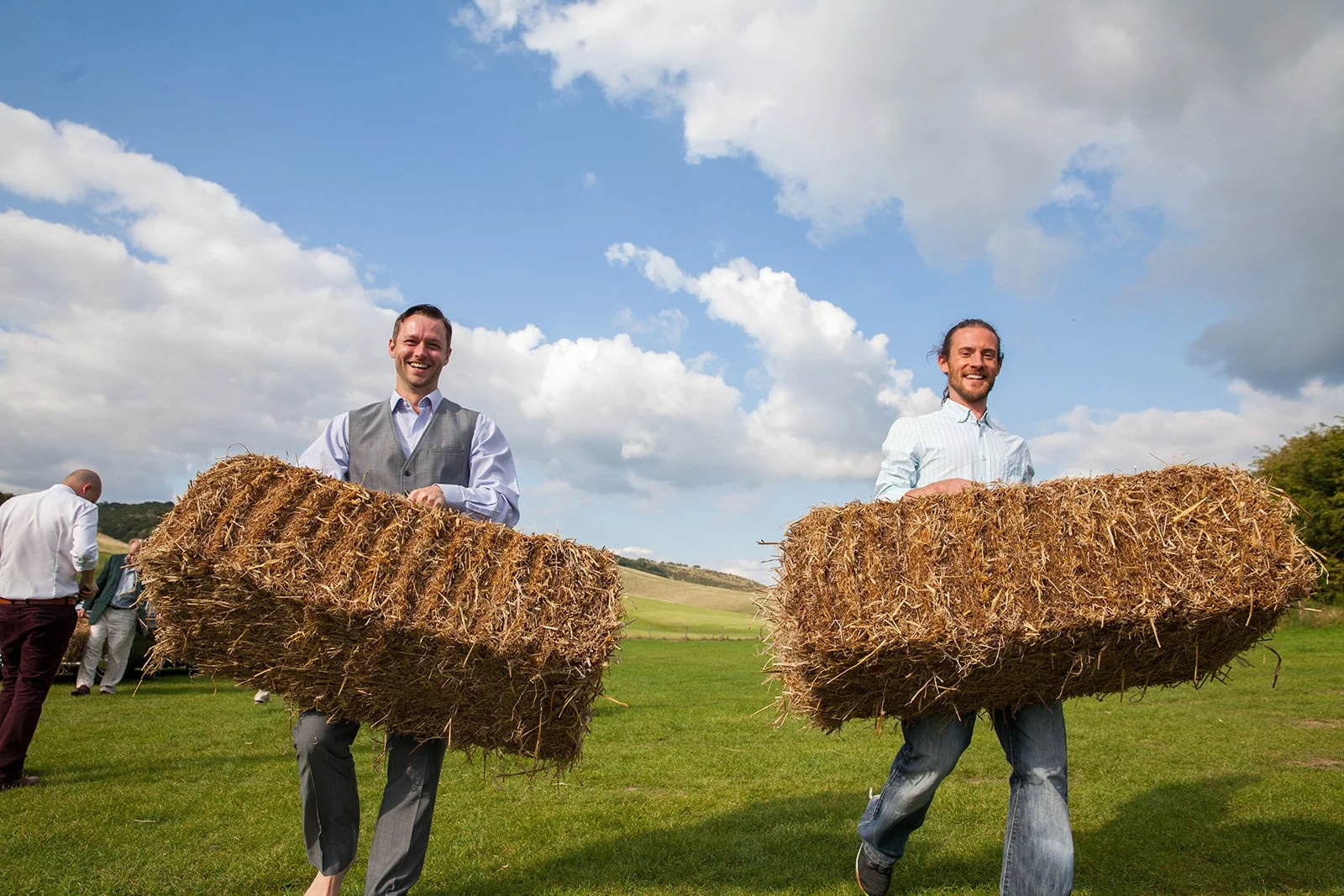 Two men in formal shirts and vests carrying hay bales across a green field under a partly cloudy sky, with other people in the background.