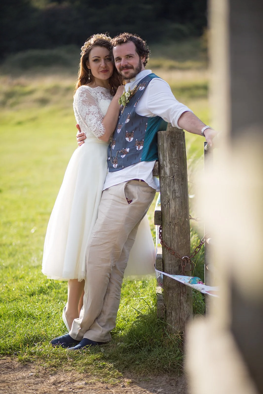 A couple in wedding attire standing outdoors next to a wooden fence, with the woman wearing a white lace wedding dress and the man wearing a white shirt and patterned vest, surrounded by a grassy field and trees.