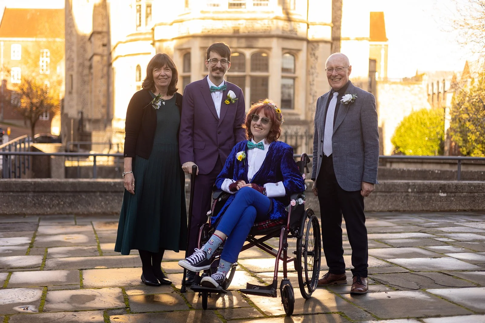 Group of five people, including a woman in a wheelchair, standing outdoors on a stone-paved area in front of a historic building during sunset. They are dressed semi-formally, some wearing floral boutonnieres and boutonnières. The woman in the wheelc