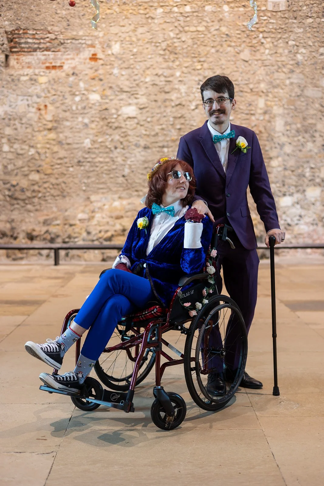 A man and a woman in a wheelchair posing together in front of an old brick wall. The woman is wearing sunglasses, a floral crown, and a blue velvet blazer, while the man is standing beside her holding a cane and wearing a suit with a bow tie and bout