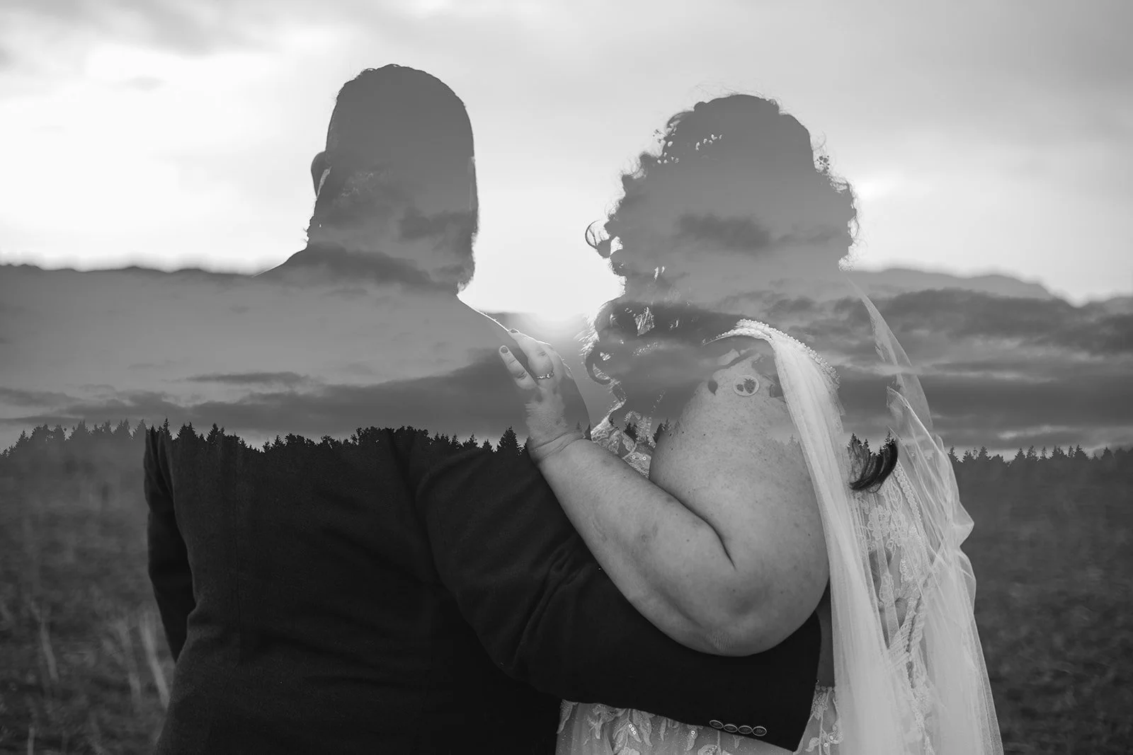 A black and white photo of a couple standing outdoors with a scenic mountain landscape in the background, captured during sunset or sunrise.