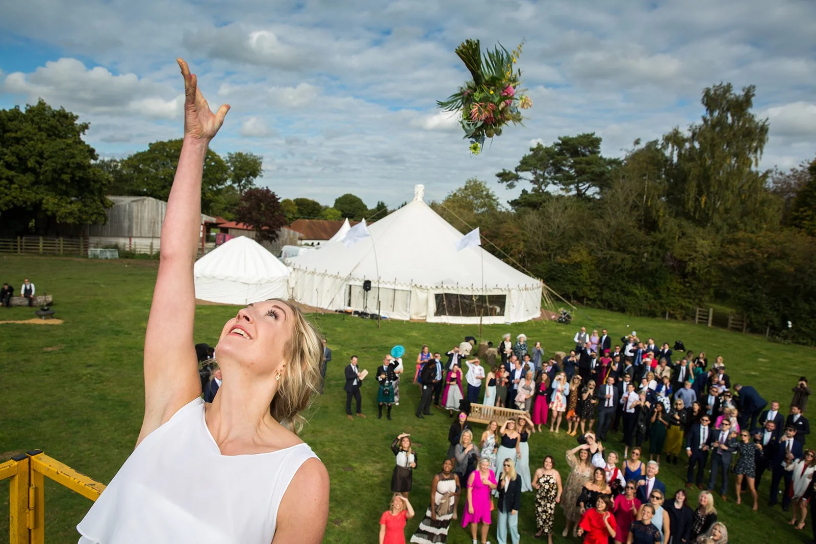 A woman tossing a bouquet of flowers at an outdoor wedding reception with a large tent and many guests in formal attire on a grassy field under a partly cloudy sky.