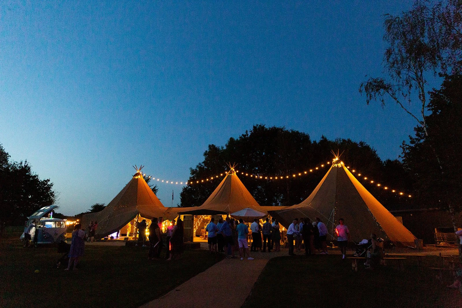 People gathered outside a large, illuminated tent at night with string lights overhead and trees in the background.