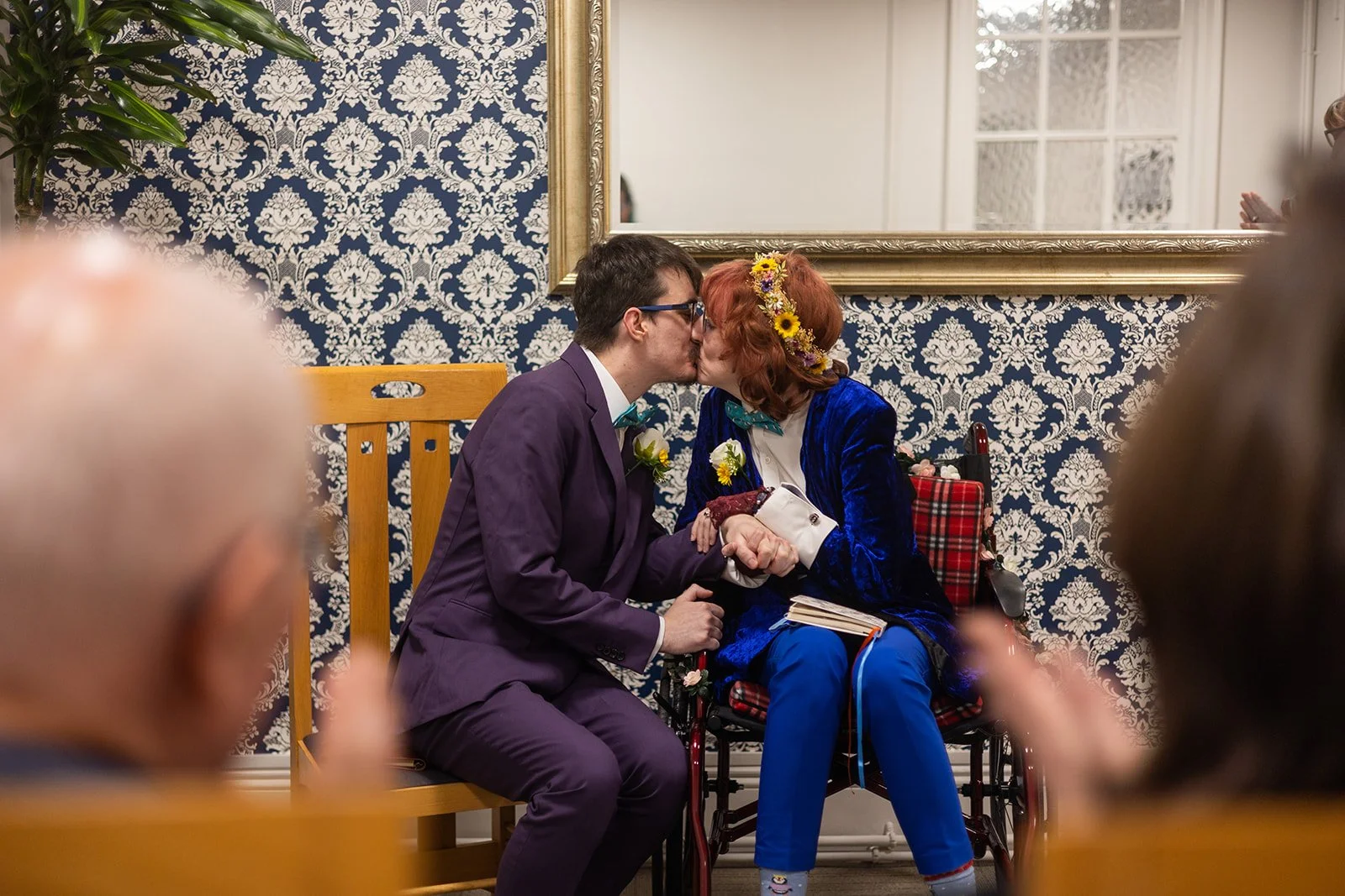 A man and a woman kissing at a wedding ceremony. The man is dressed in a purple suit with glasses and a bow tie, and the woman is in a blue velvet jacket with a floral headband. The woman is seated in a wheelchair, holding a book, and the man is knee